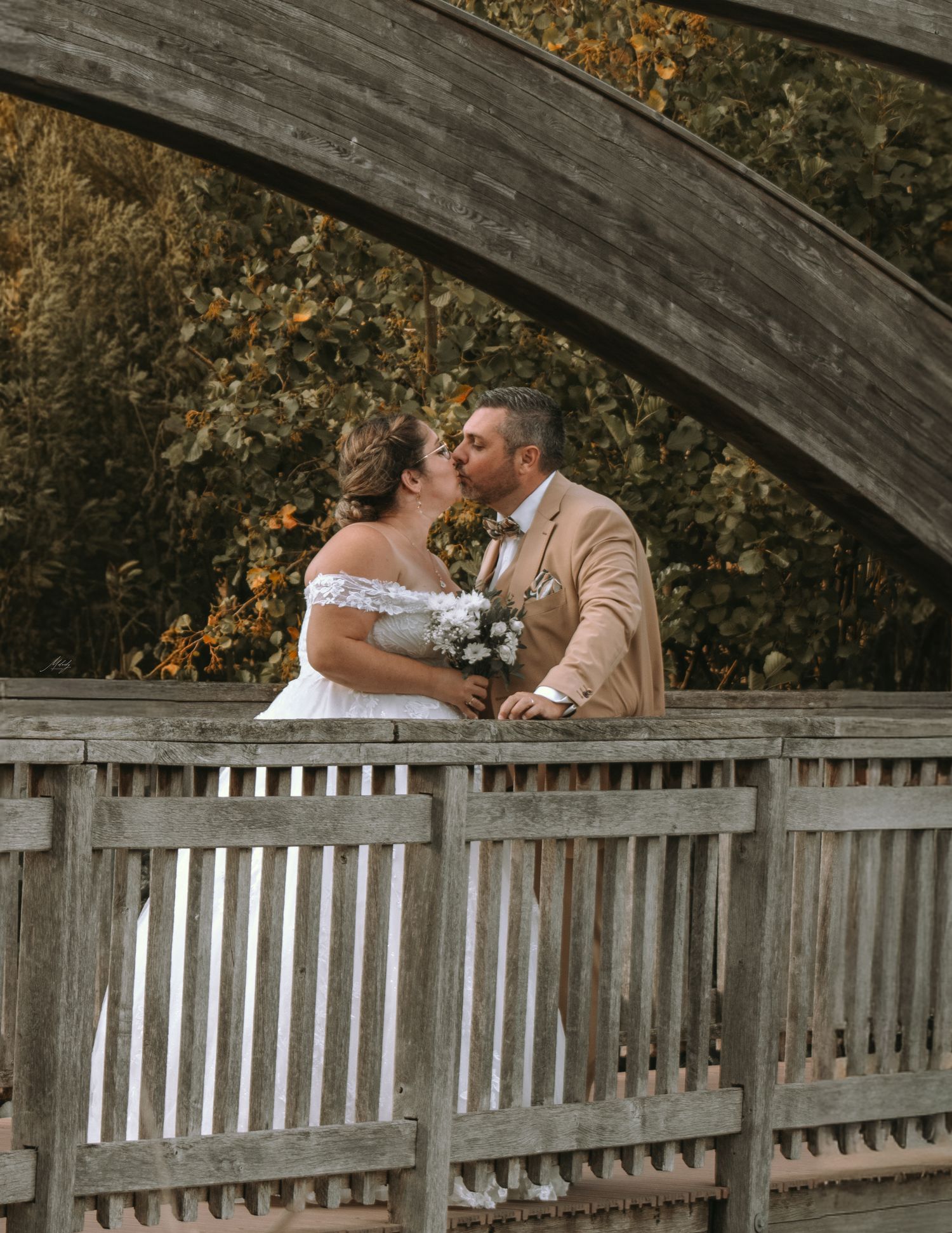 Bride and groom kissing on a wooden bridge framed by a curved arch with lush autumn foliage.