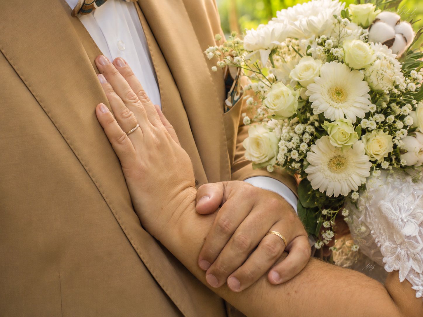Close-up of bride and groom holding hands showing wedding ring beside a white floral bouquet.