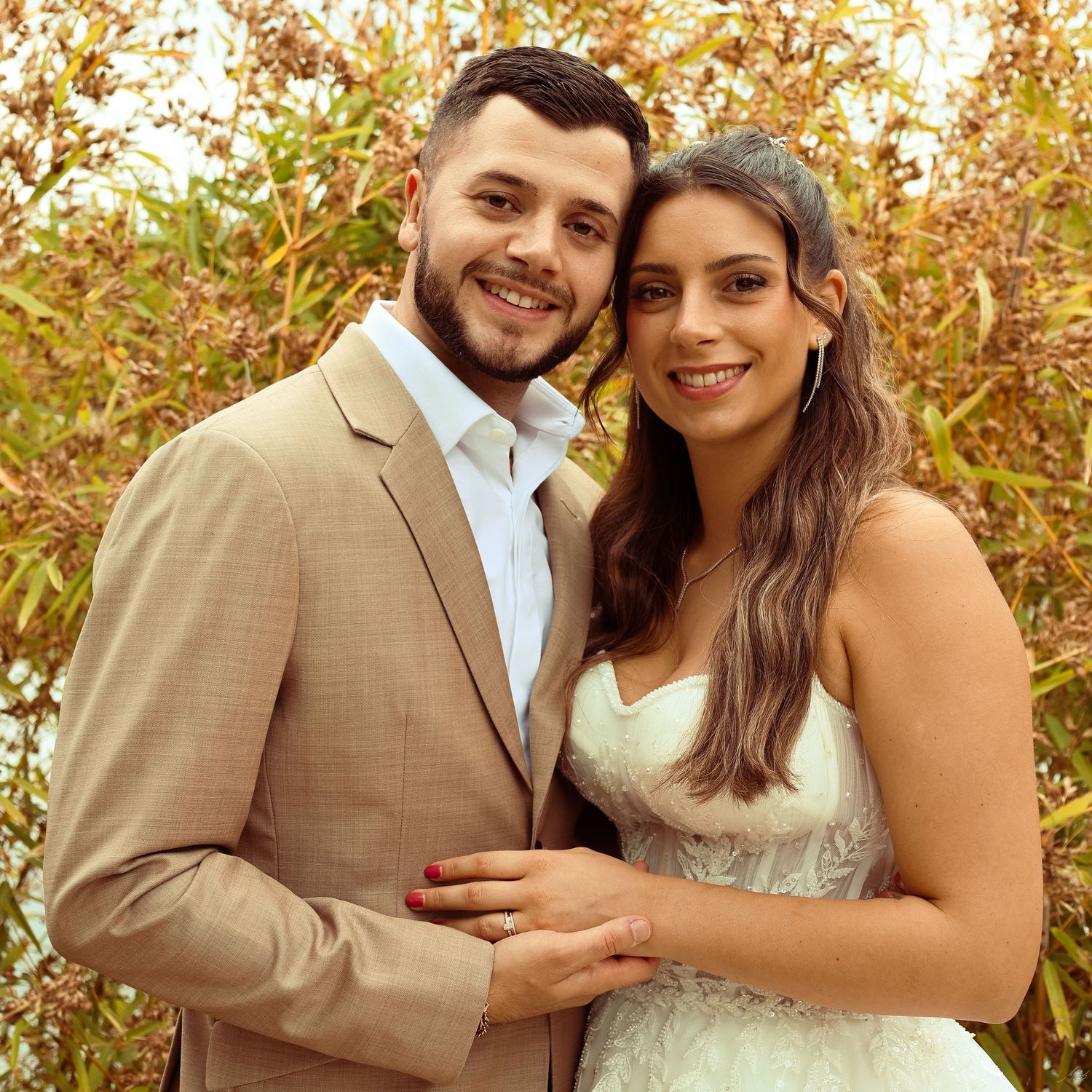 Smiling bride and groom posing together outdoors, groom in beige suit and bride in white dress.