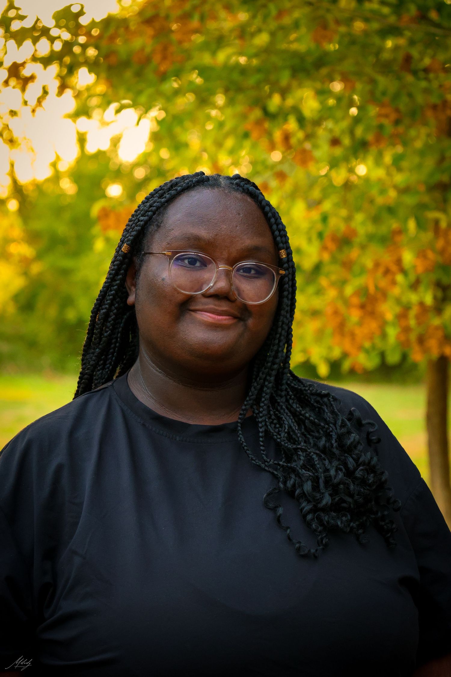 Young woman with braids and glasses smiling outdoors in front of colorful autumn foliage.