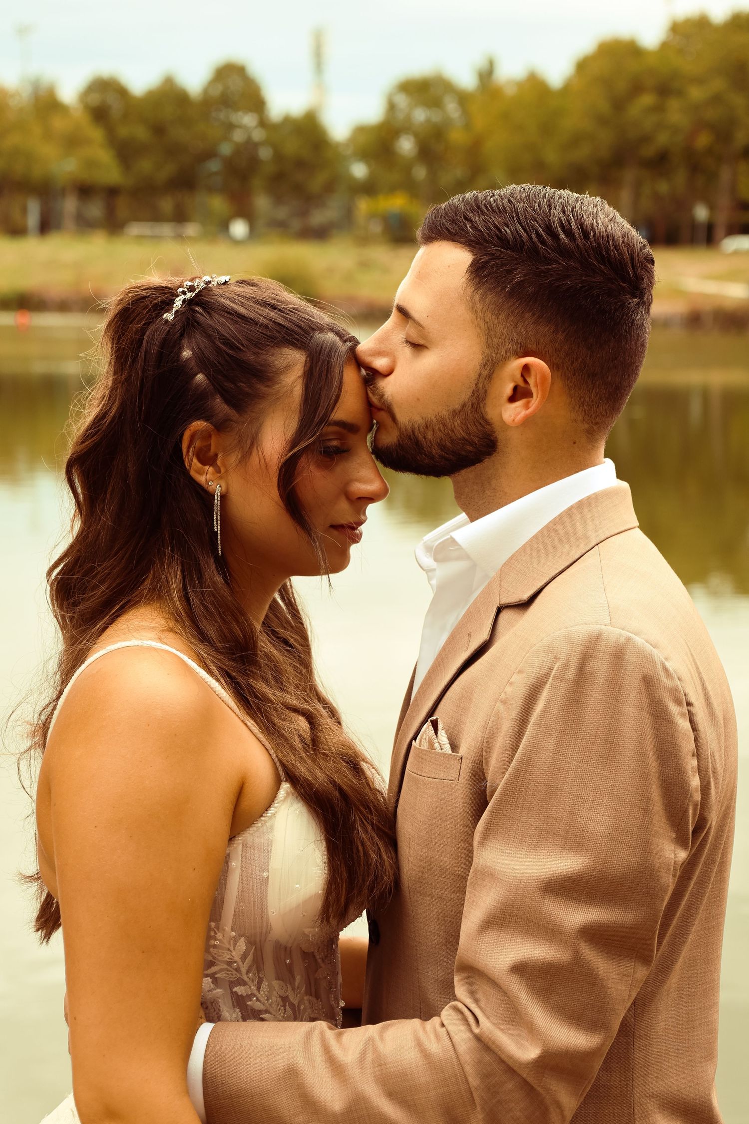 Groom tenderly kisses bride on forehead near a serene lake, both dressed in elegant wedding attire.