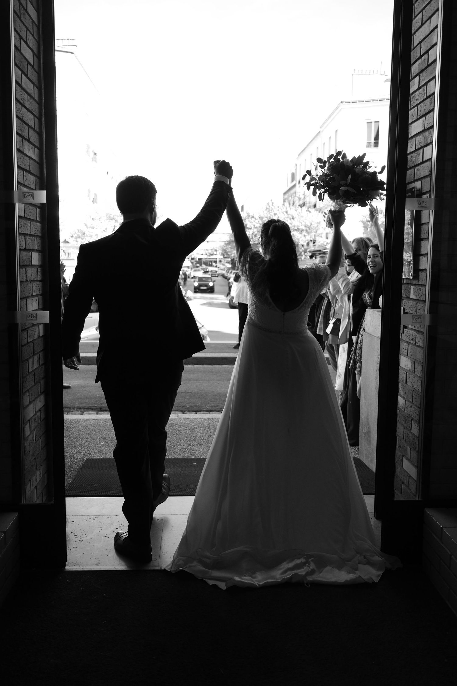 Silhouette of bride and groom exiting church doors with raised hands and bouquet in black and white.
