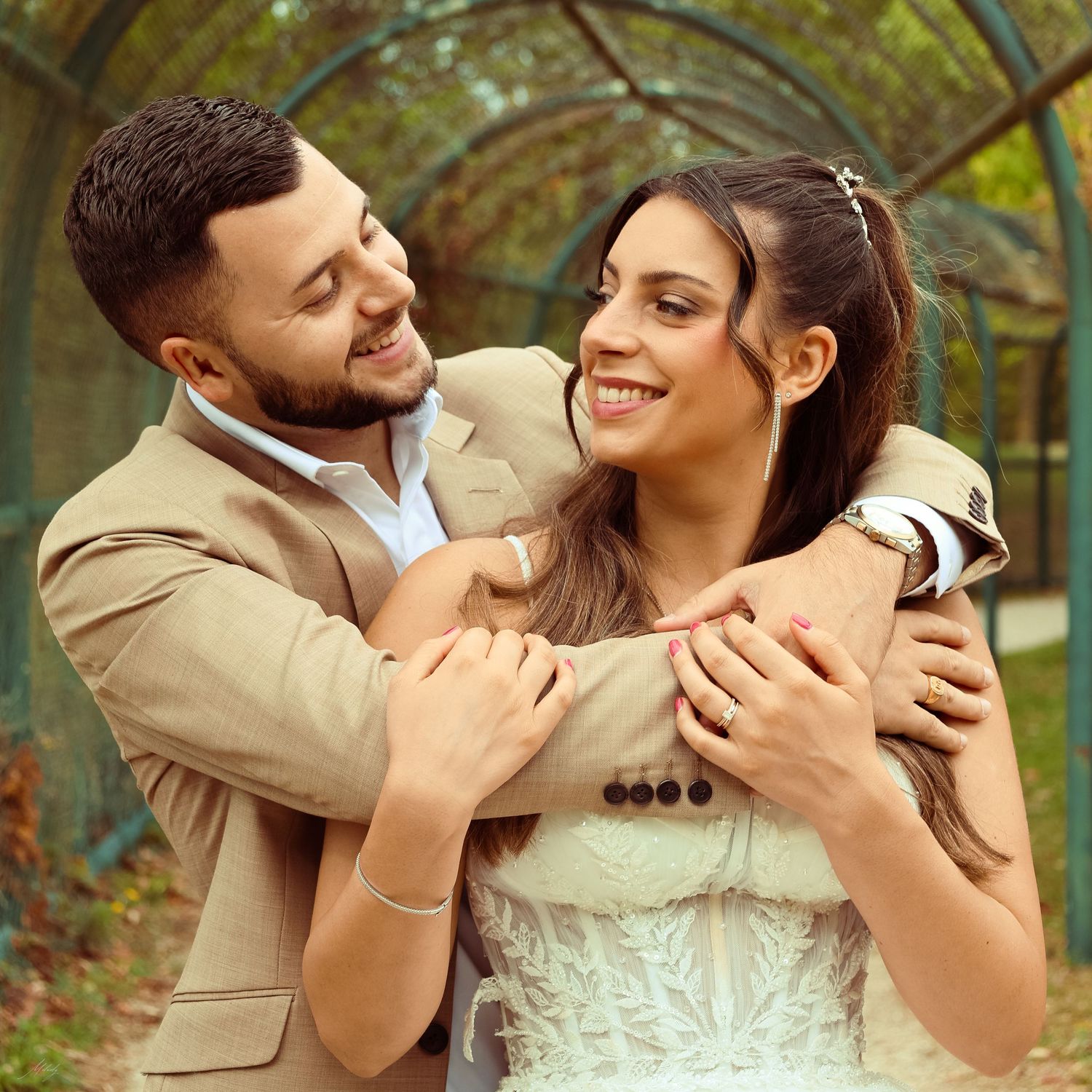 Groom embracing bride from behind as both smile joyfully under a green garden archway.