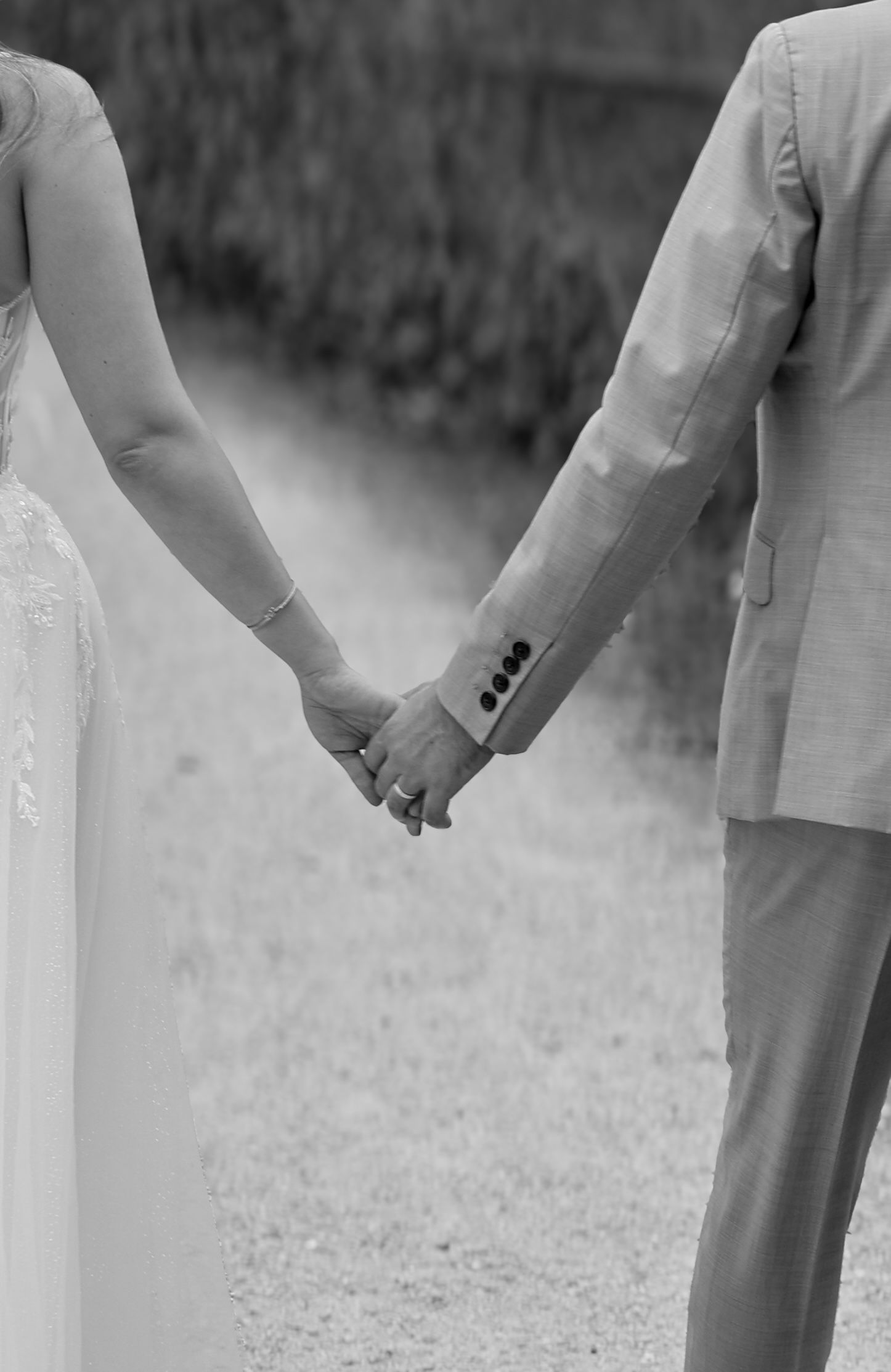 Black and white photo of bride and groom holding hands while walking down a garden path.