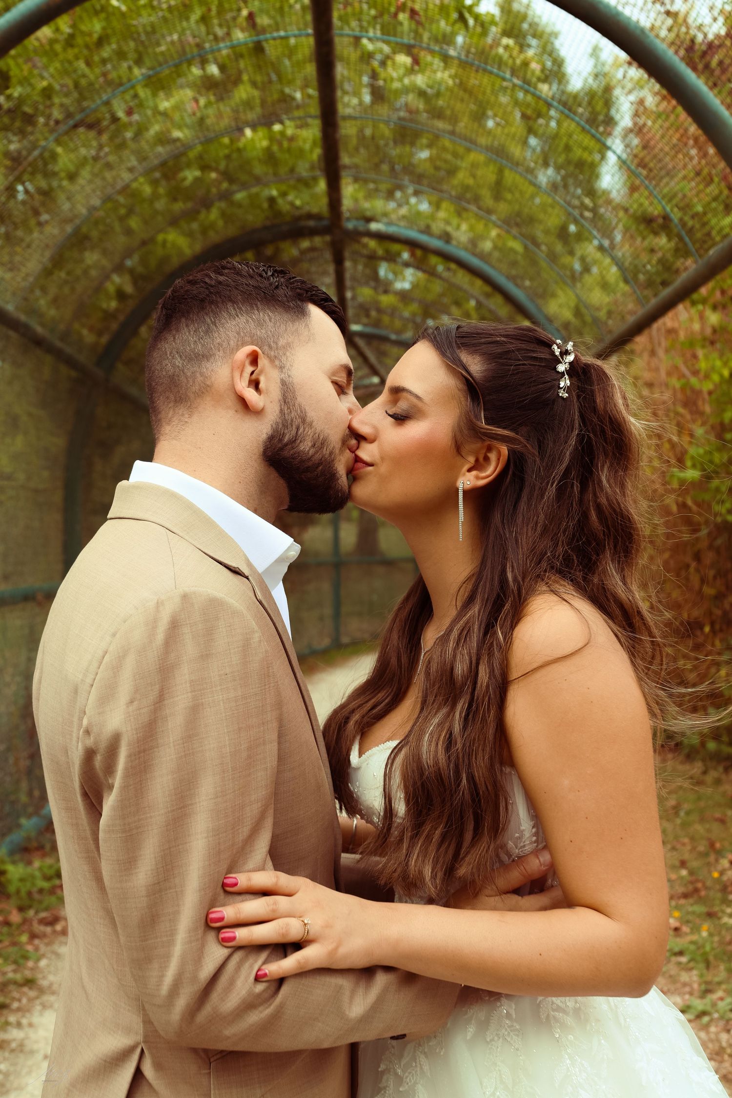 Bride and groom sharing a passionate kiss inside a green tunnel archway on their wedding day.