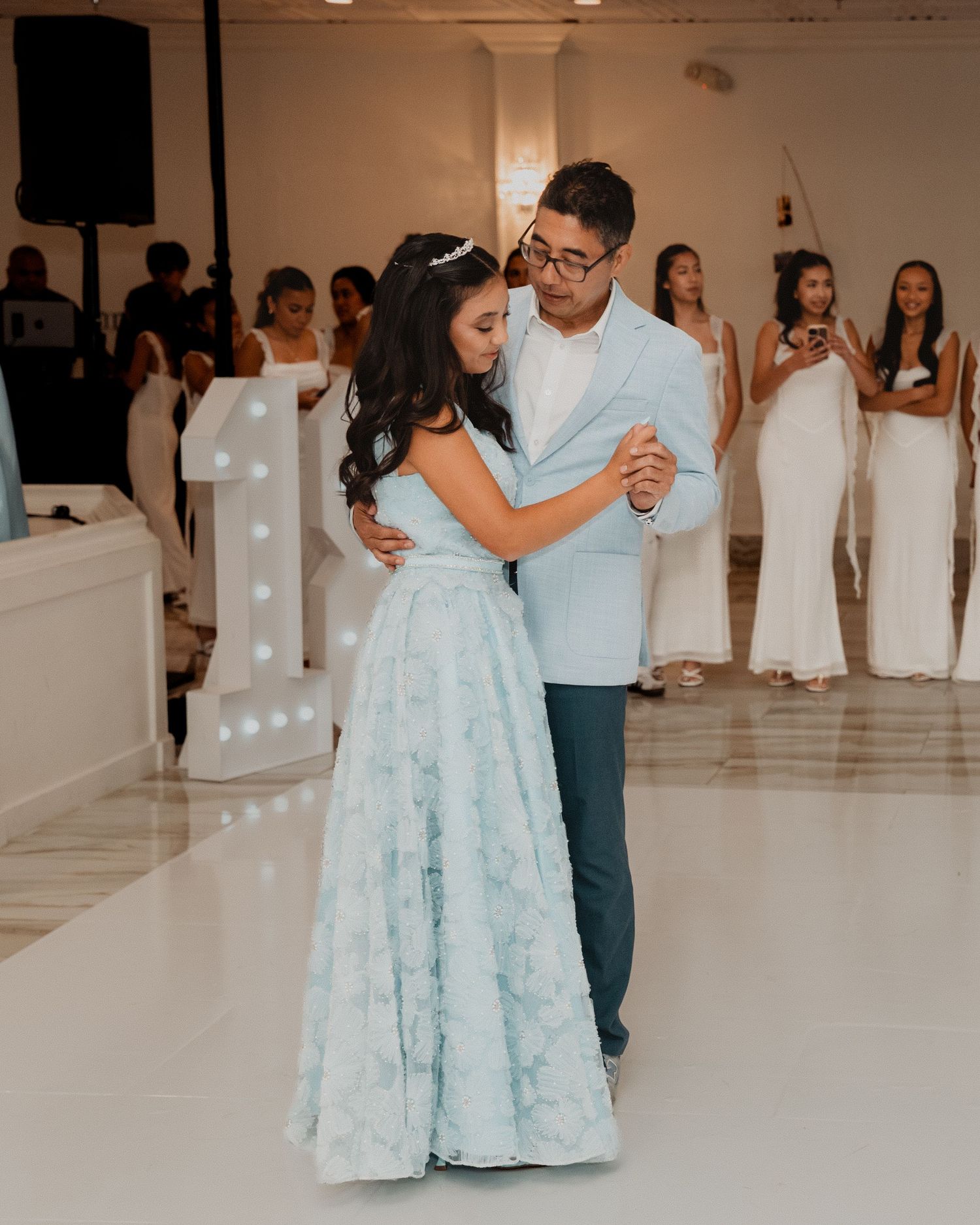 A young woman in a light blue floral gown dances with a man in a blue suit at a quinceañera celebration.