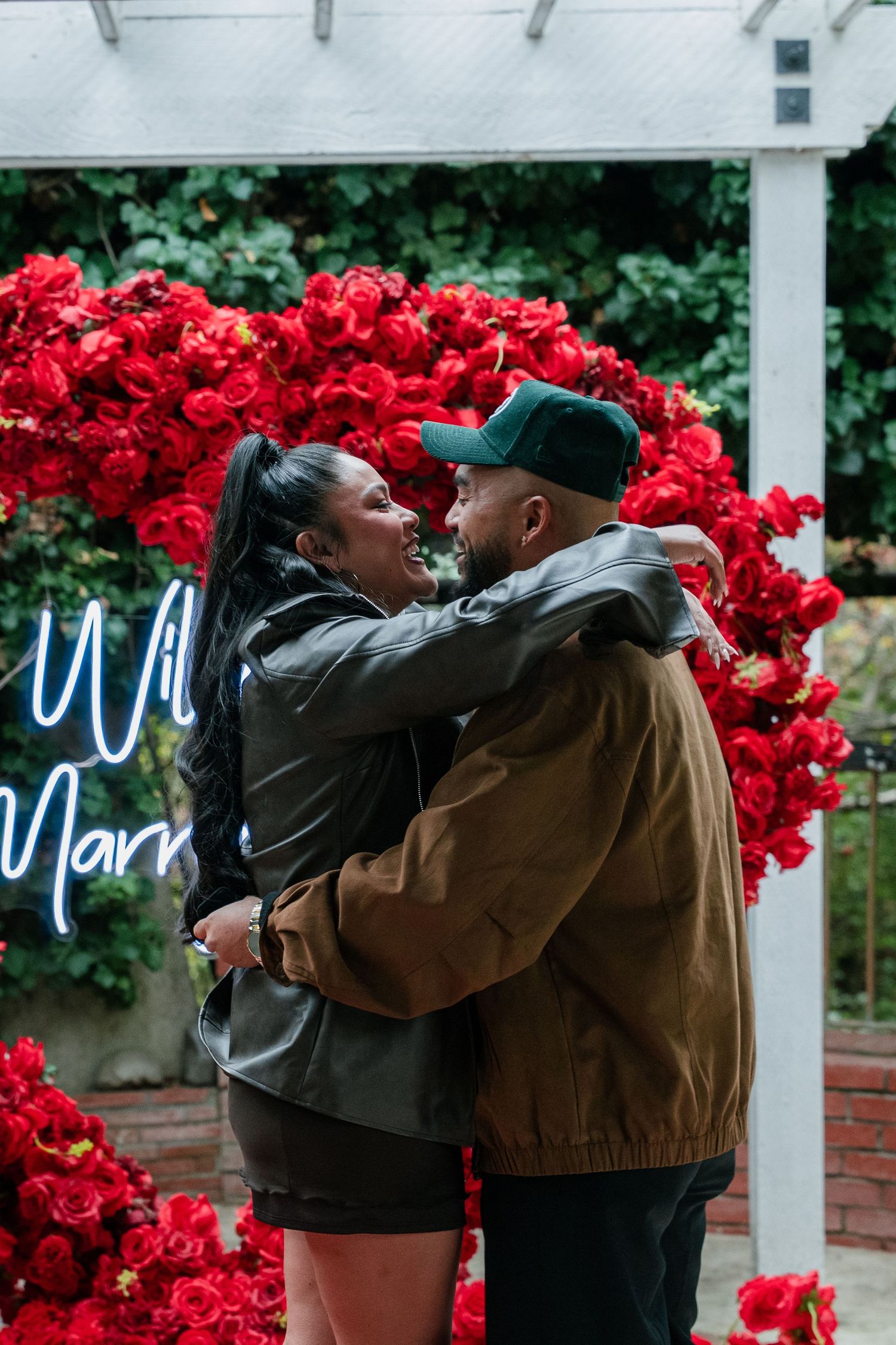 Two people in brown jackets share an intimate moment against a backdrop of vibrant red flowers and white pillars.