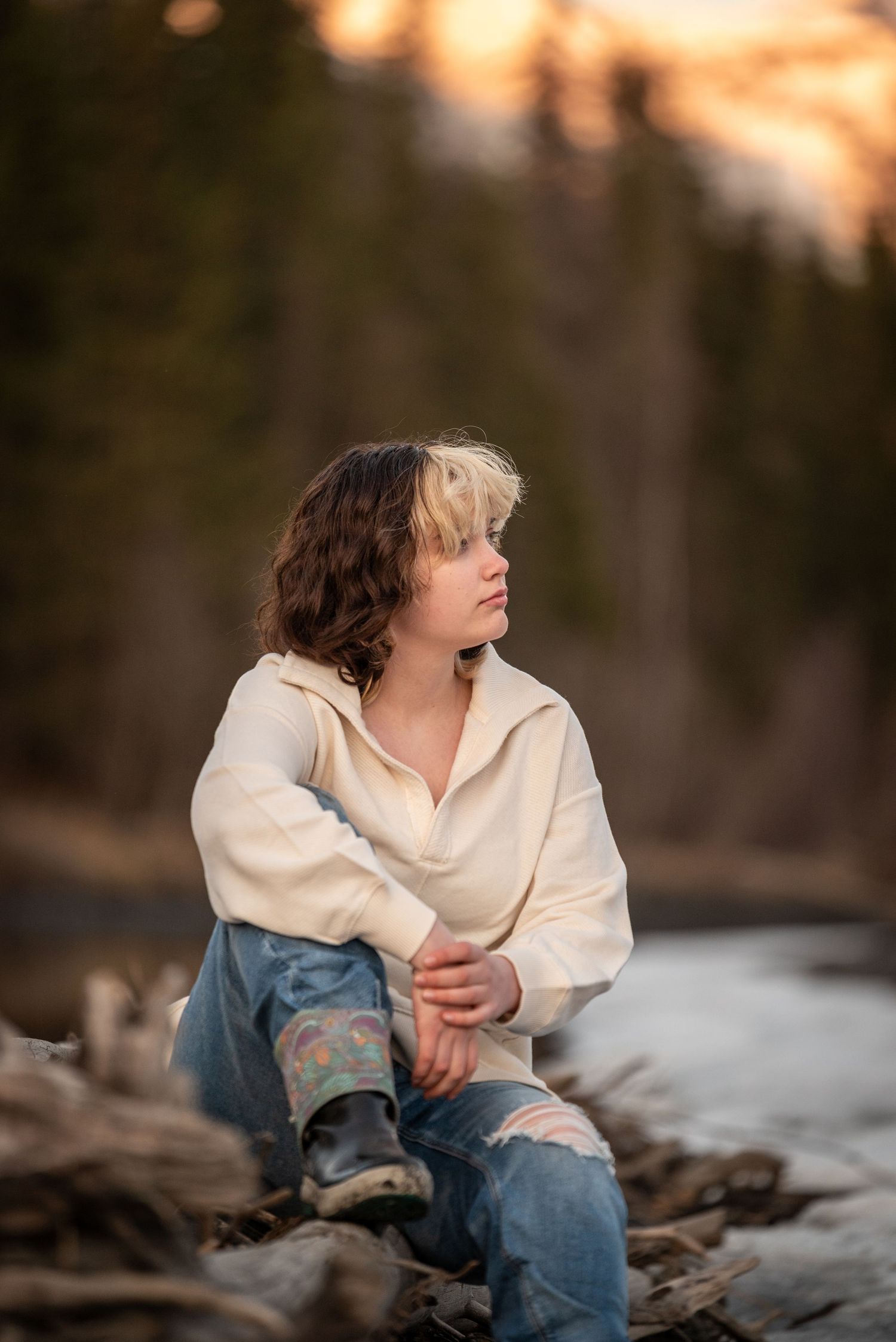 Person in cream sweater and ripped jeans sits thoughtfully on rocky shore during golden sunset.