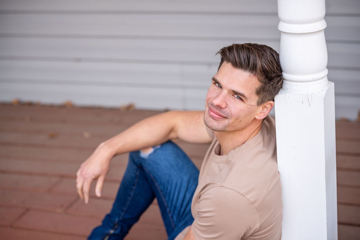 A person in a tan shirt and blue jeans sits casually on white stairs while leaning against a pillar.