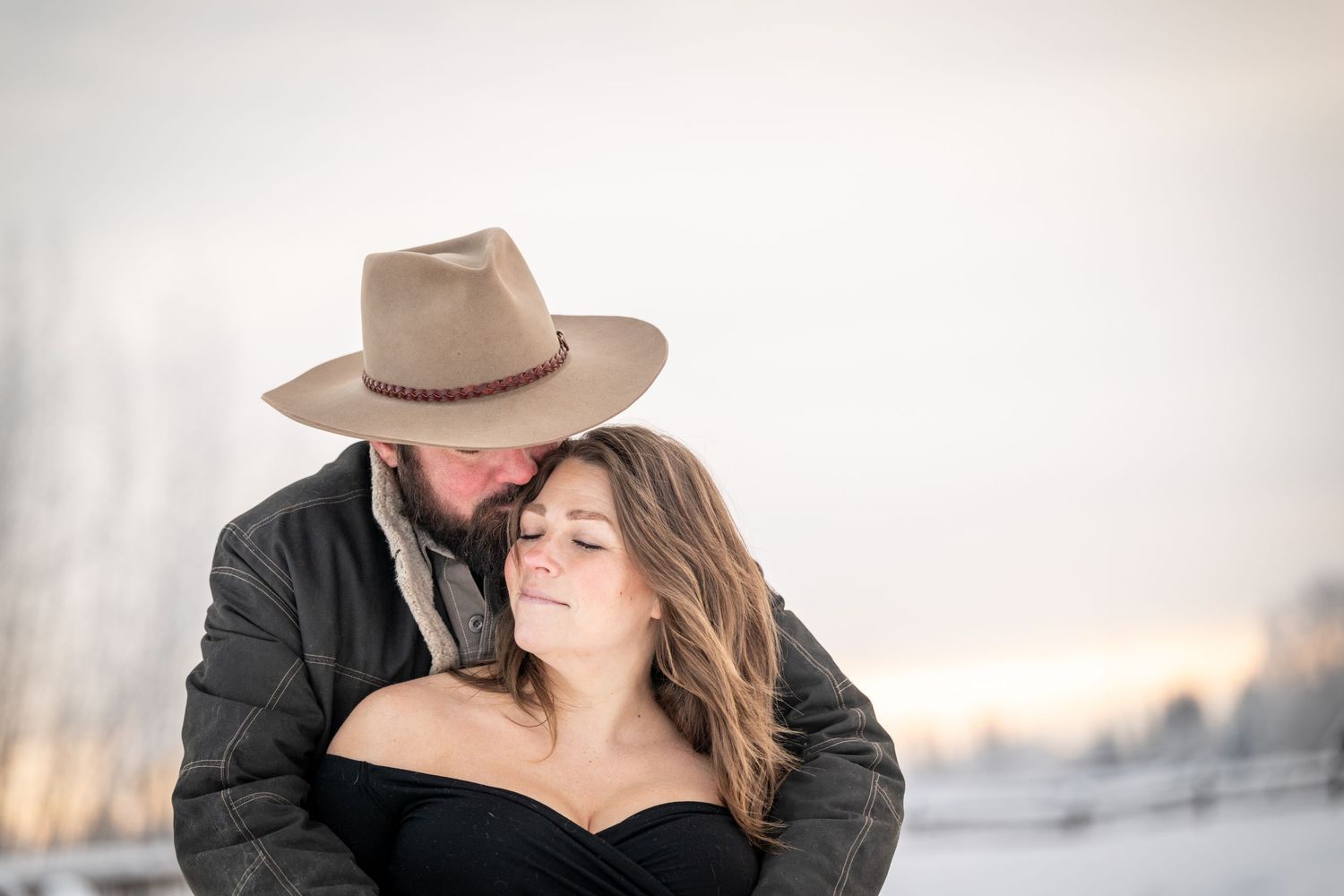 A romantic couple embraces in a soft, dreamy outdoor setting with winter light and neutral tones in the background.