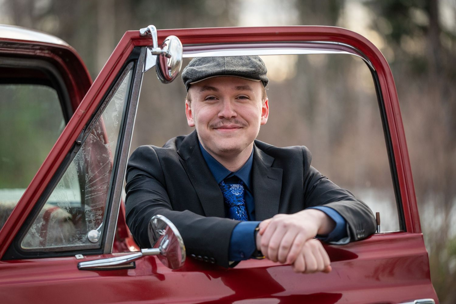 Smiling young man in flat cap and blazer leaning out of classic red truck window outdoors.
