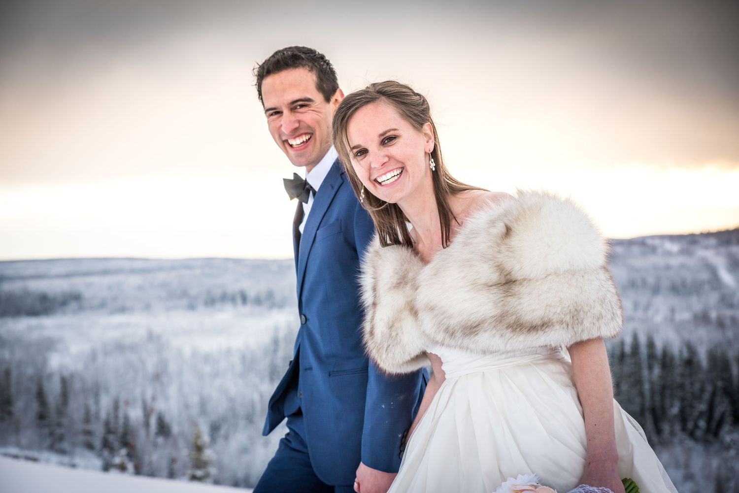 A couple in winter wedding attire poses together in a snowy mountain landscape with trees in the background.  Whitney McLaren Photography
