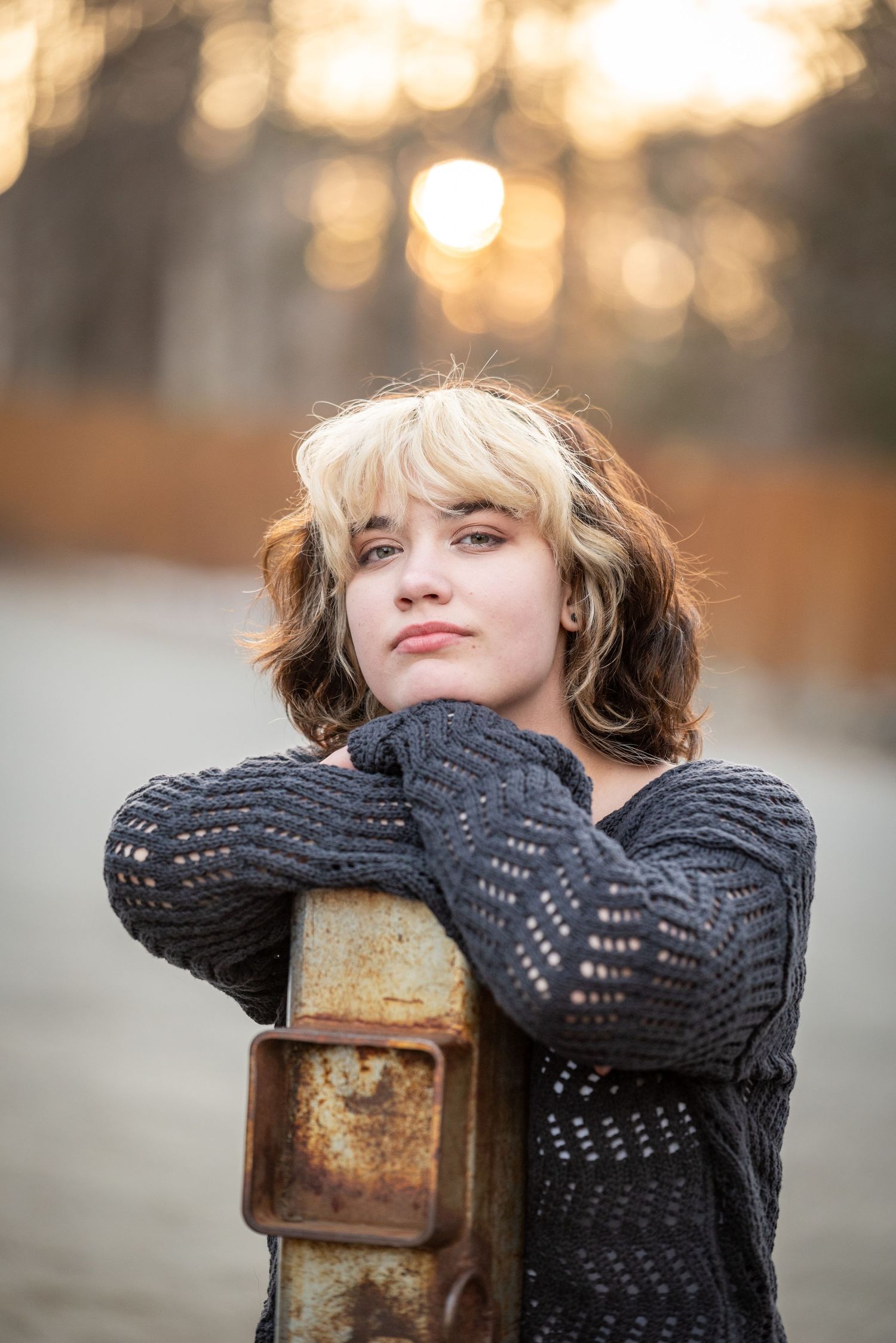 Young woman in gray knit sweater leans on rusty post at sunset in a serene outdoor setting.