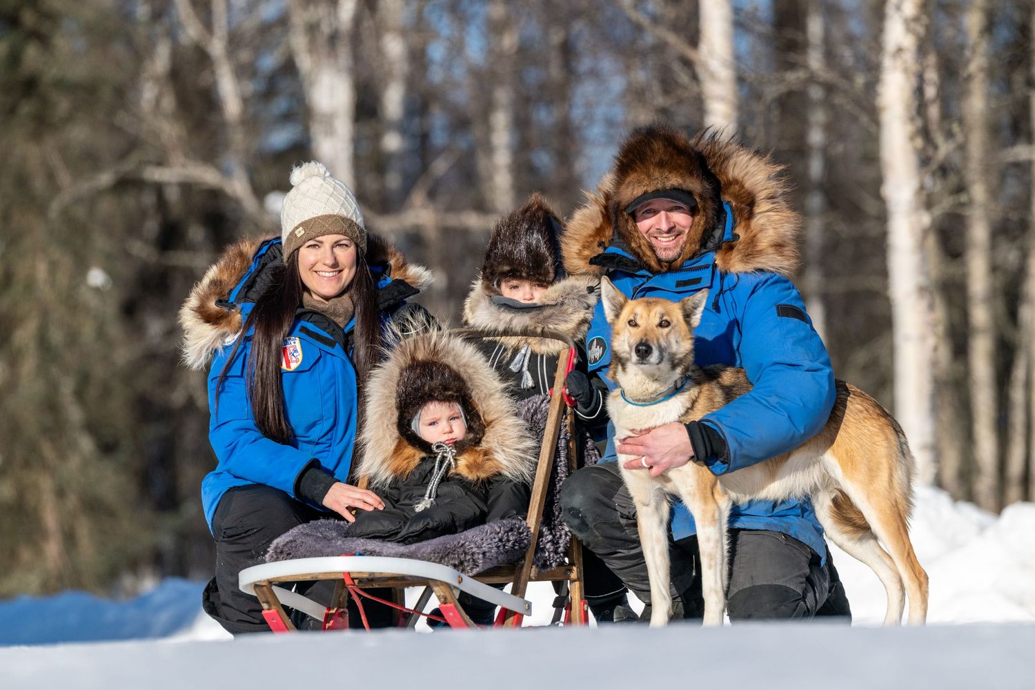 Family in blue winter jackets posing with sled dogs in snowy forest, smiling outdoors in bright winter sunshine.