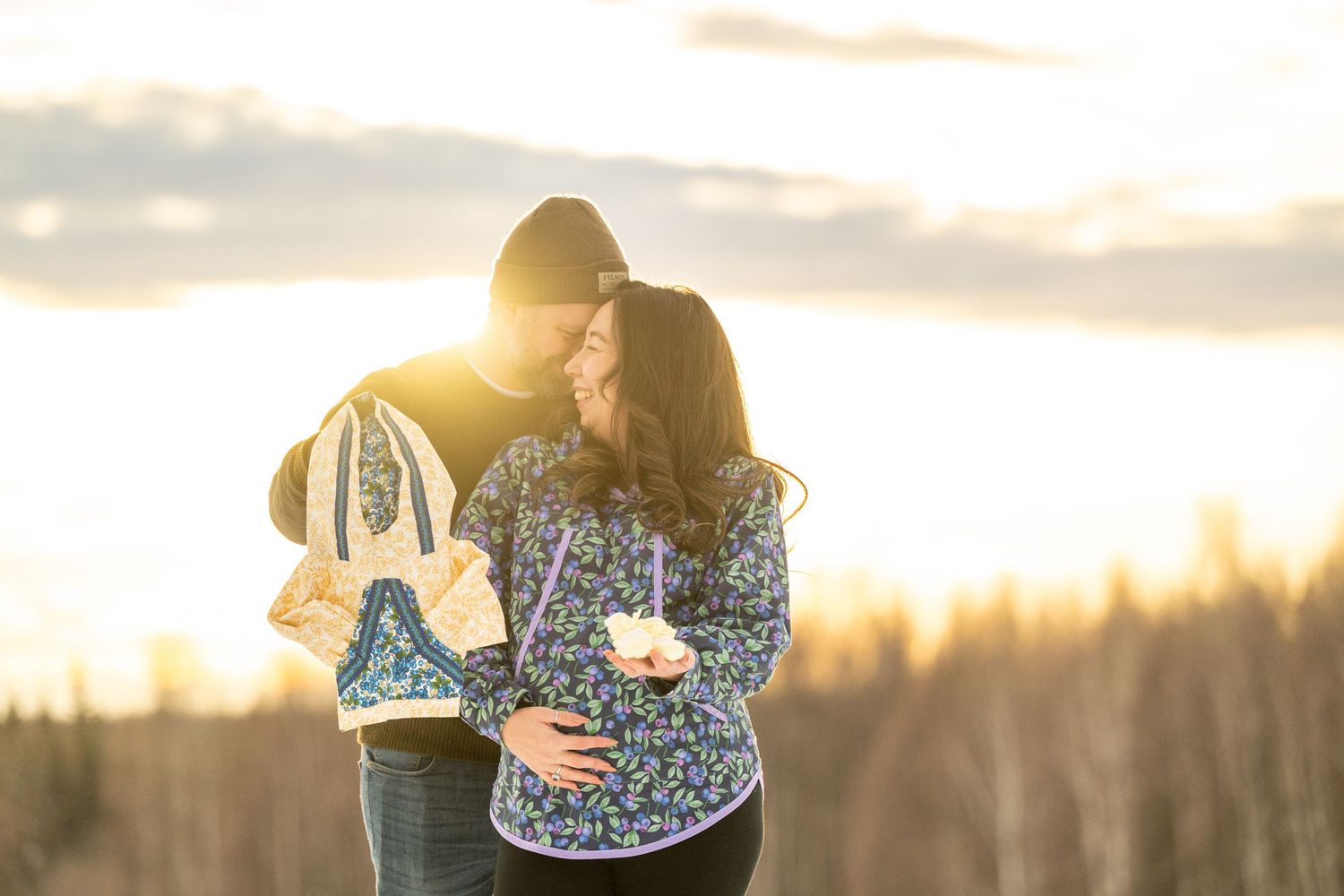 Couple embracing at golden sunset outdoors, man holding hockey skates, woman in floral jacket, trees in background.