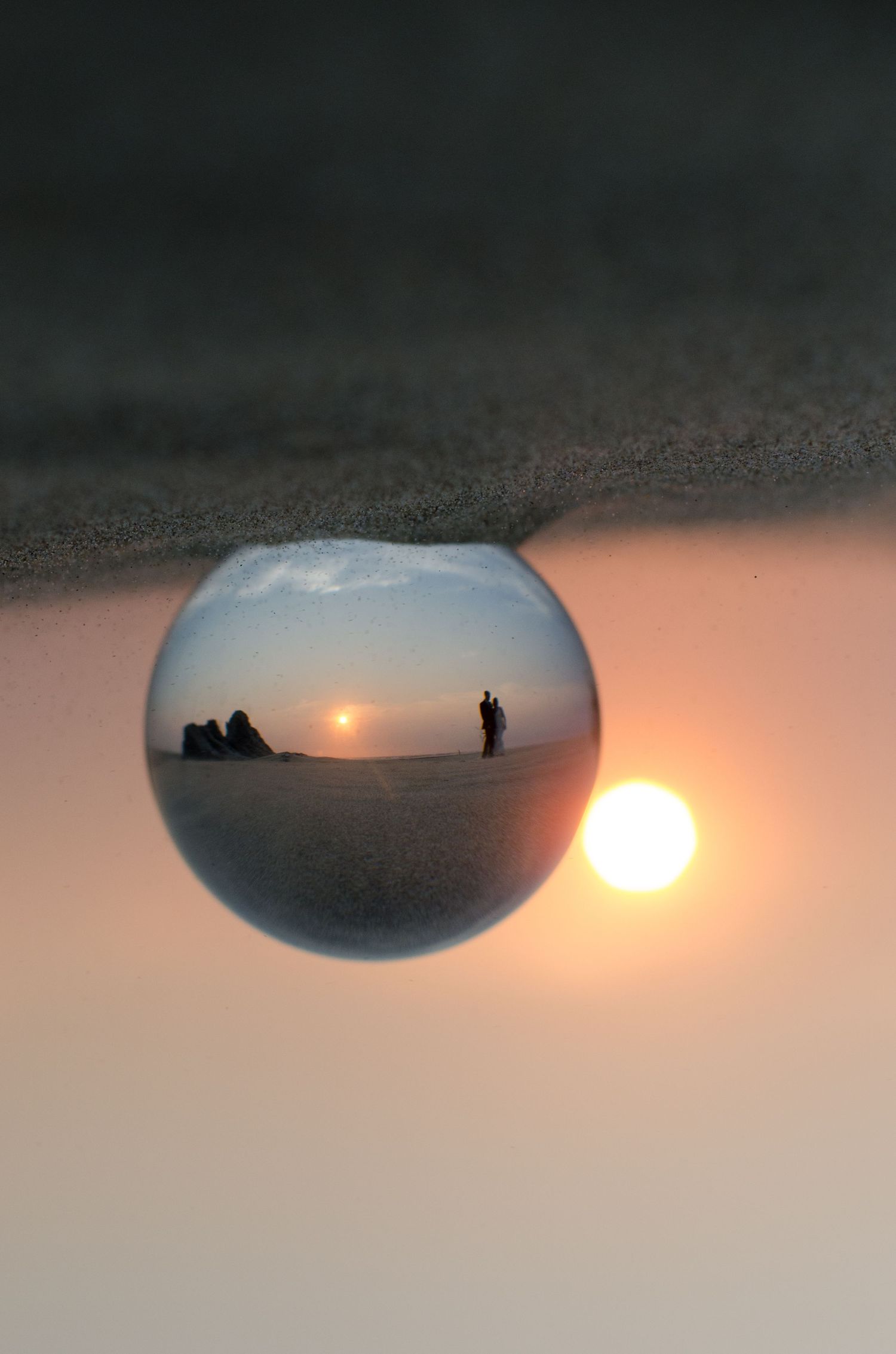 A silhouetted figure stands against a sunset sky reflected in a small handheld mirror on a dark surface.