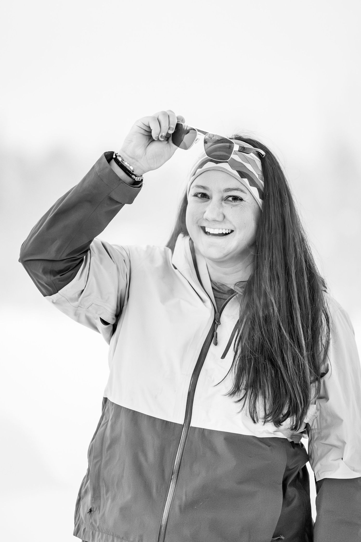 Smiling young woman in ski jacket lifting sunglasses on snowy day, black and white winter sport portrait.