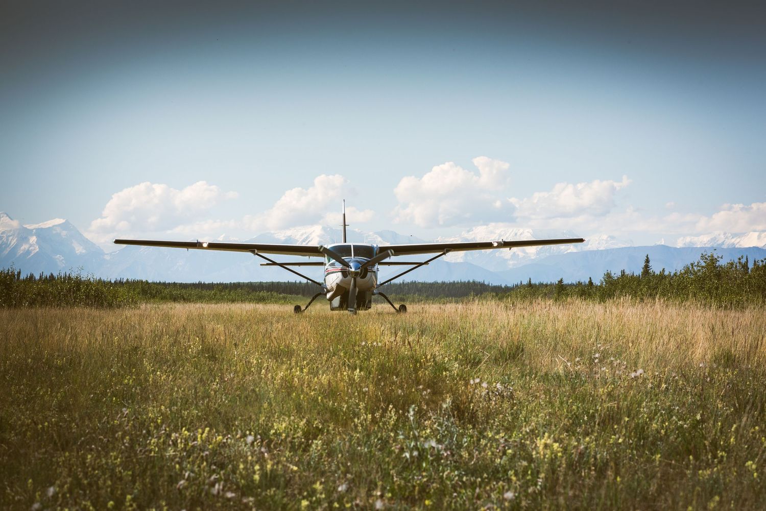A small propeller plane parked on a grassy airstrip with mountains and blue sky in the background.