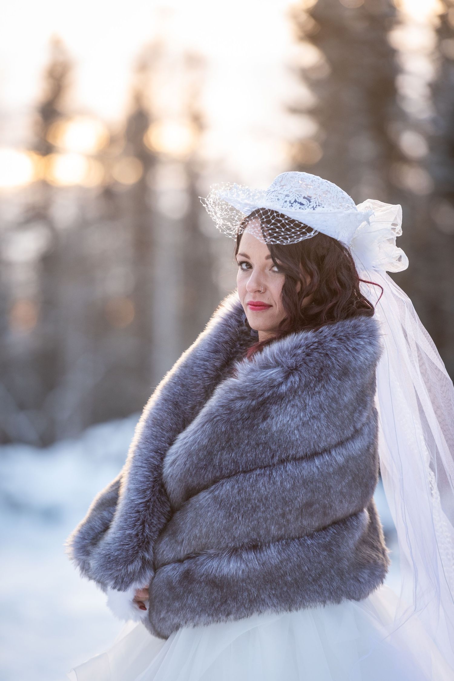 Winter bride wearing grey fur stole and veil against snowy forest backdrop. Whitney McLaren Photography