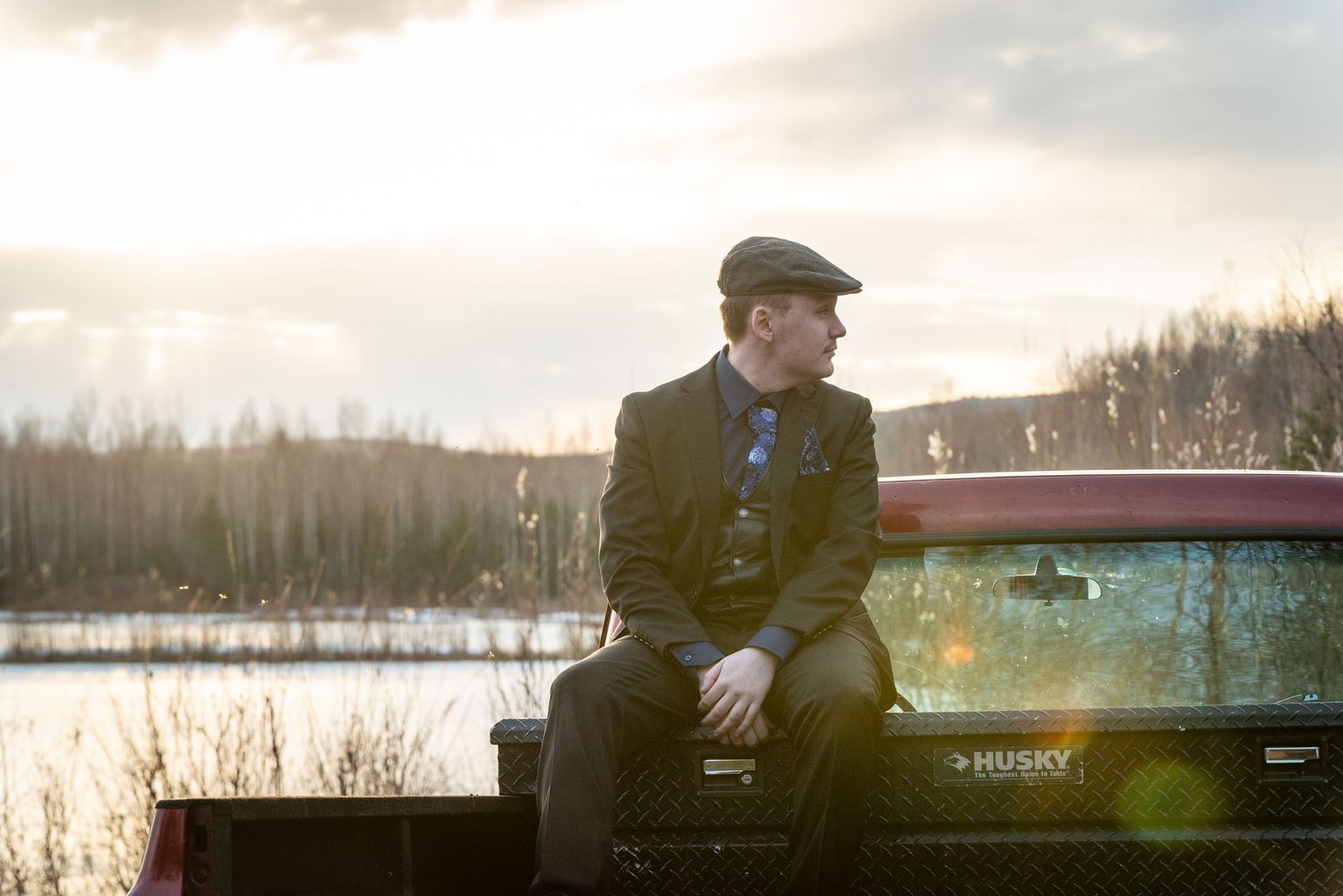Man in flat cap sitting on red truck tailgate by a lake at sunset in a scenic outdoor setting.