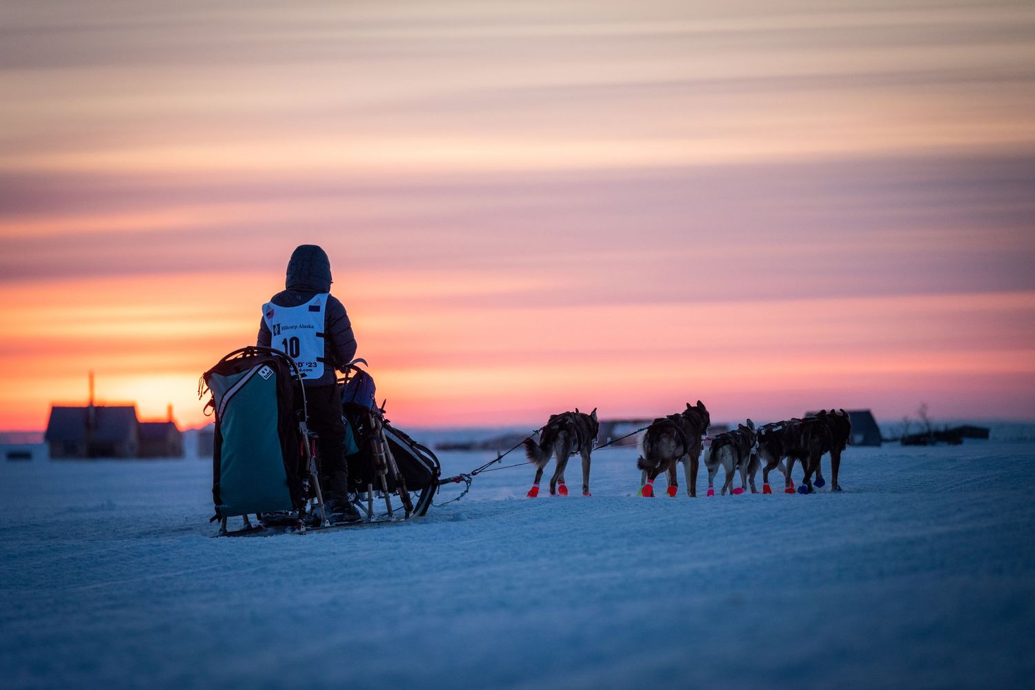 Dog sleds traverse across a frozen landscape during a vibrant sunset. Mushingphotos.com Whitney McLaren Photography