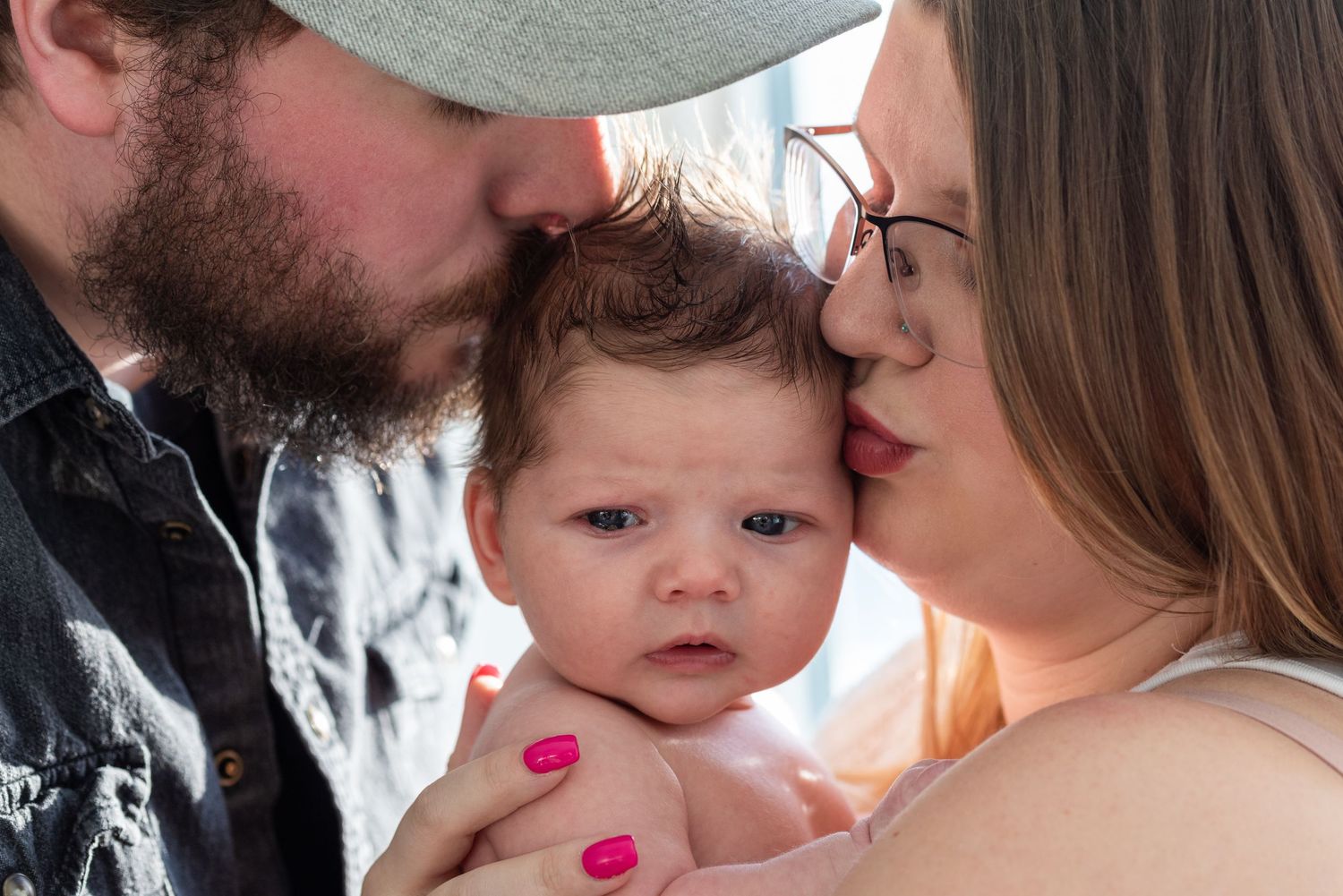 Two parents lovingly kissing their newborn baby on the head, sharing a tender family moment outdoors.