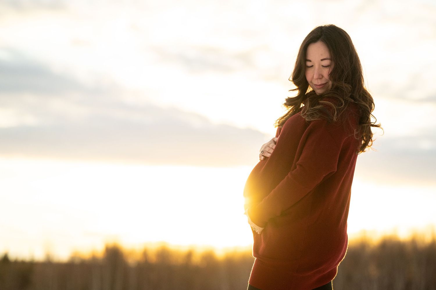 Pregnant woman in red dress cradling baby bump at golden sunset, long dark hair, serene outdoor setting.