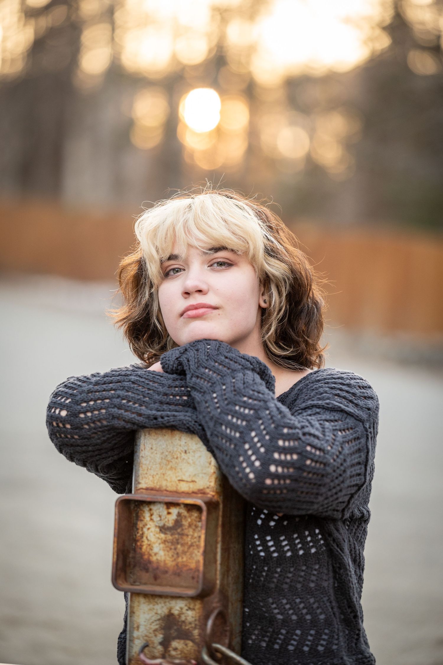 Person in dark knit sweater rests on rustic metal post with warm sunset bokeh background.