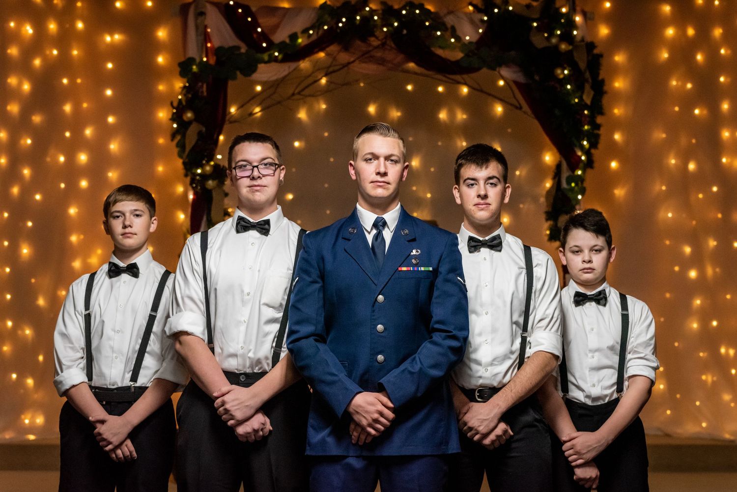 Row of young attendants in formal wear standing against a backdrop of twinkle lights. Whitney McLaren Photography