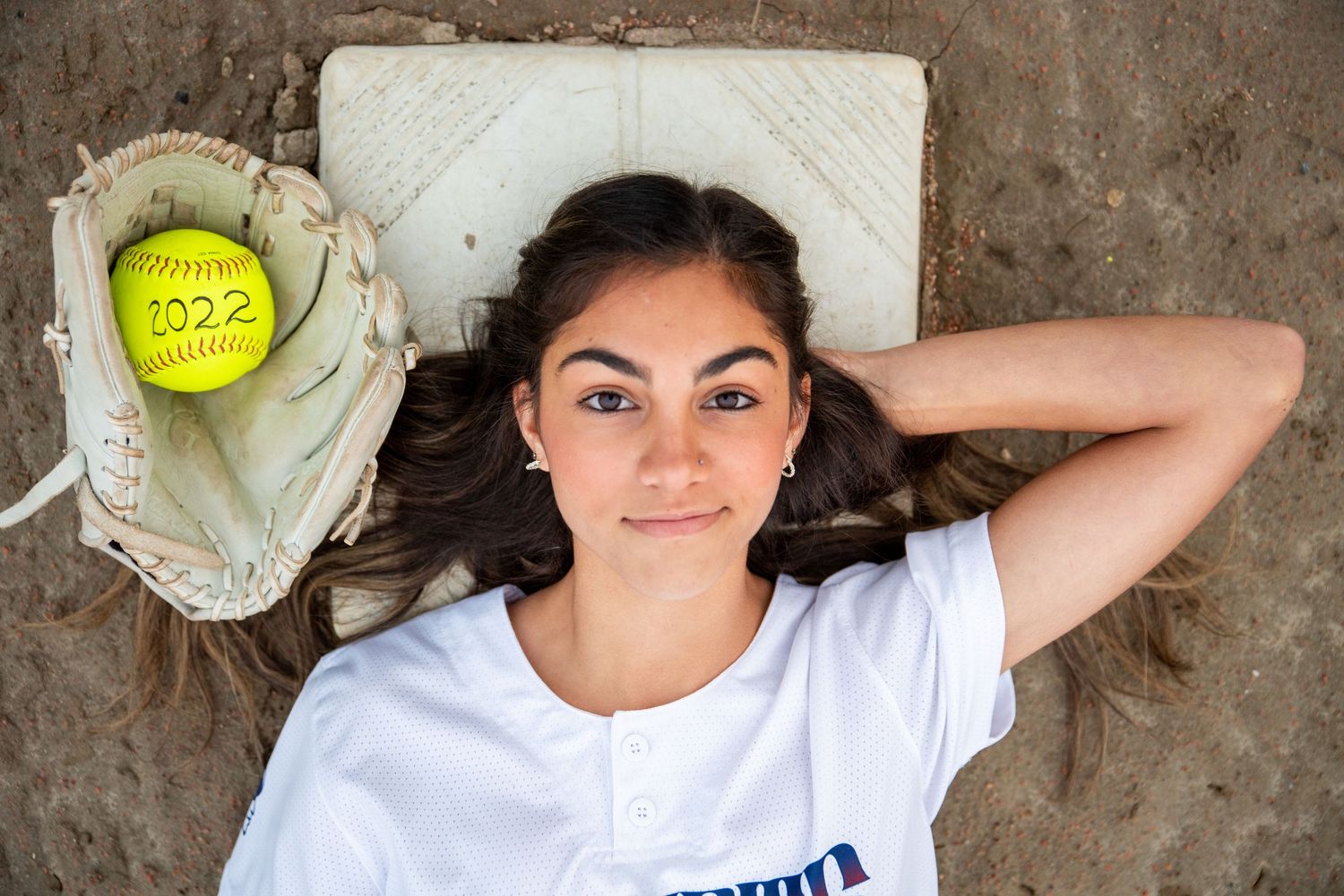 A softball player lies on the ground with a glove holding a yellow ball marked 2022, wearing a white jersey.