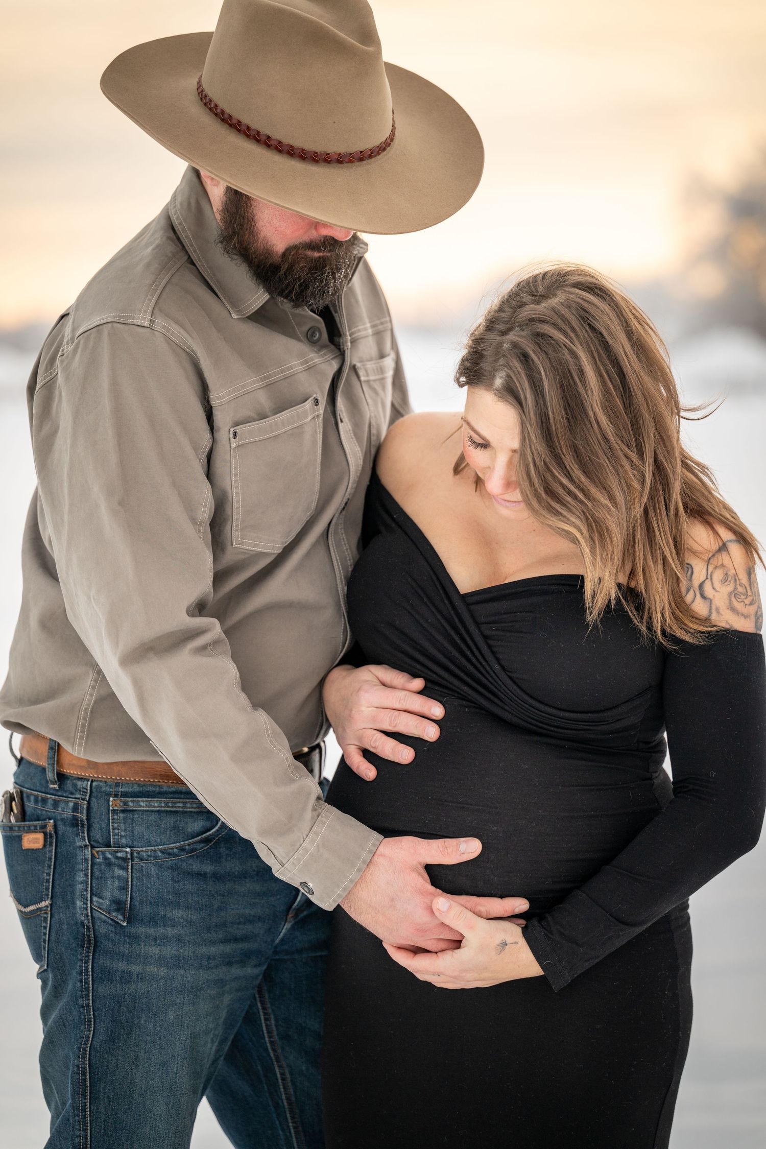 A bearded man in a cowboy hat and pregnant woman in a black dress share a tender maternity photo at sunset.