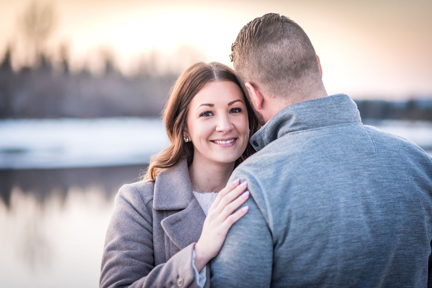 A smiling woman in a gray coat embraces a man outdoors near a frozen lake at sunset.