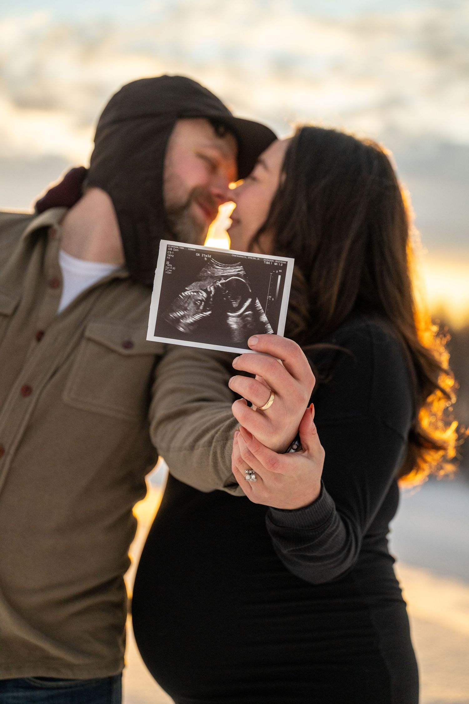 Couple kissing at sunset while holding ultrasound photo, pregnancy announcement with golden hour backdrop.