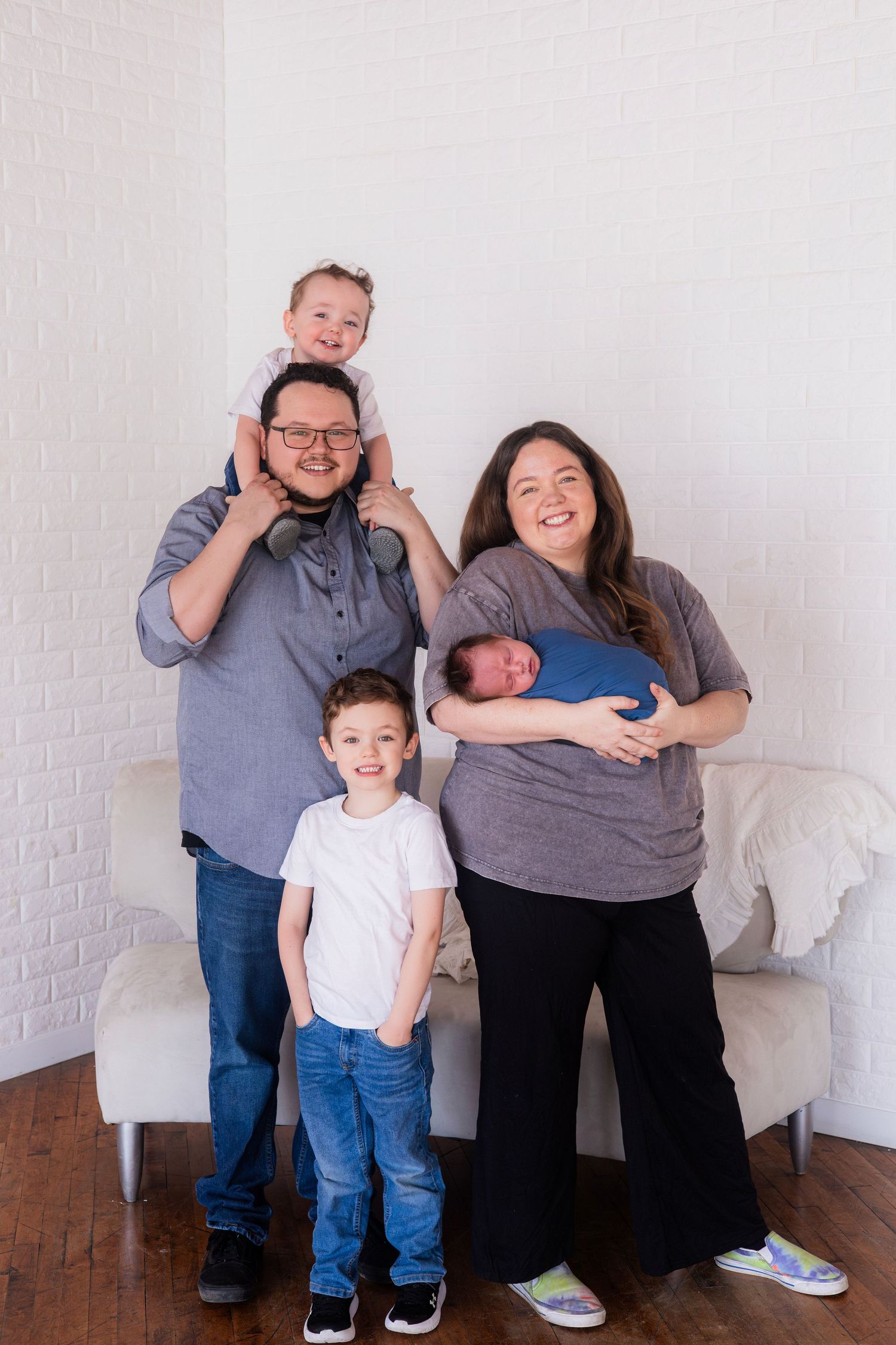 Happy family of five posing with newborn baby, father holding toddler on shoulders, mother cradling infant.