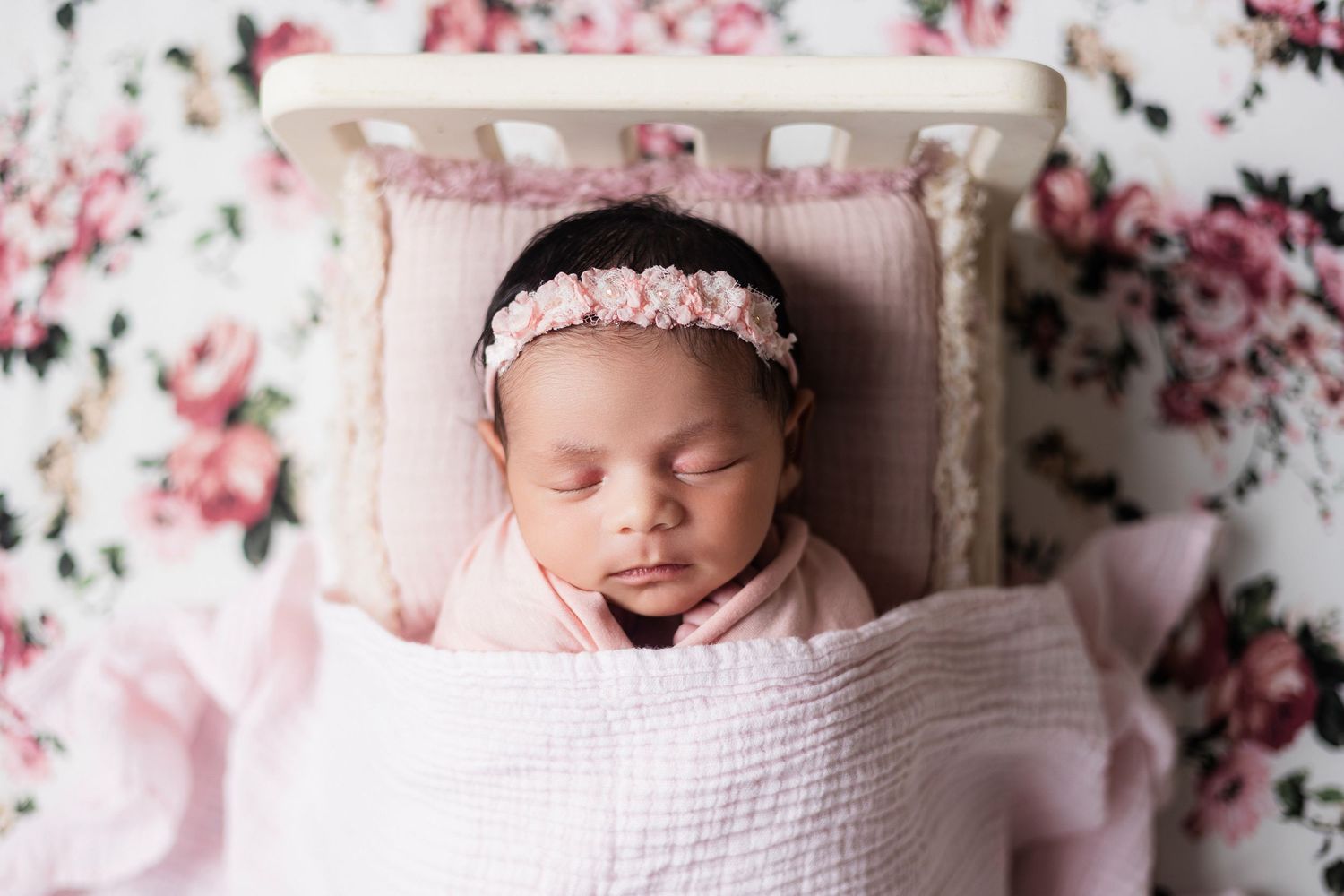 Sleeping newborn baby wrapped in pink swaddle on floral background with flower headband.