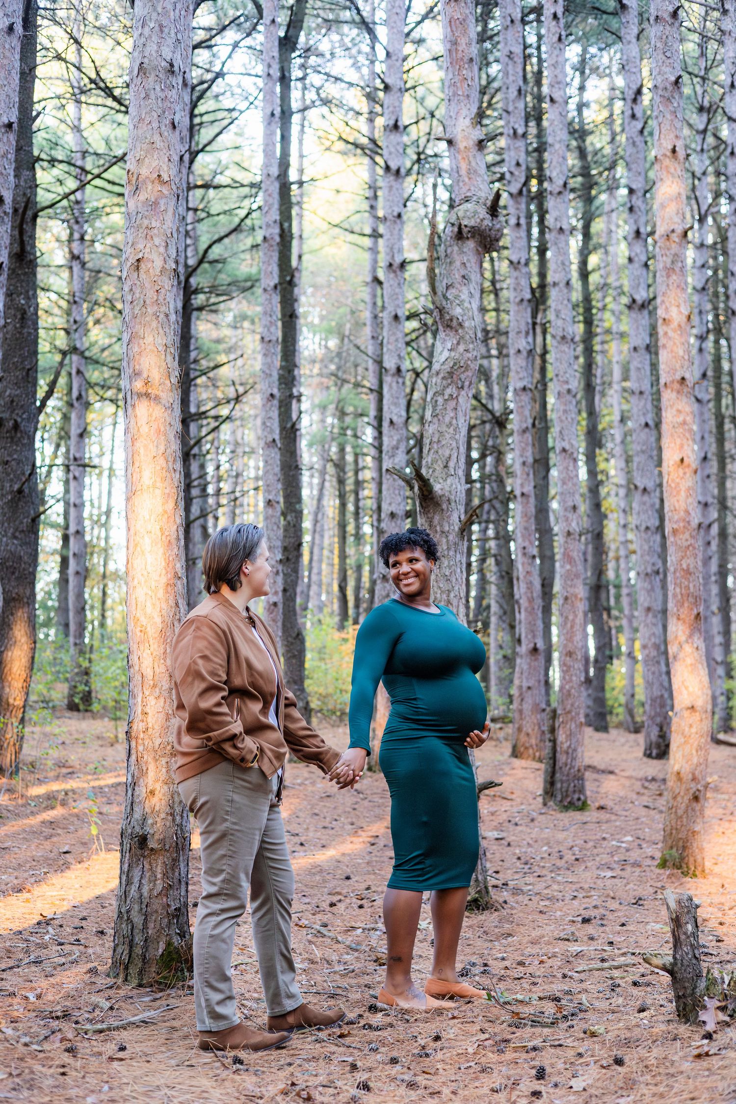 Pregnant woman in teal dress holds hands with partner in forest setting, smiling joyfully among tall pine trees.