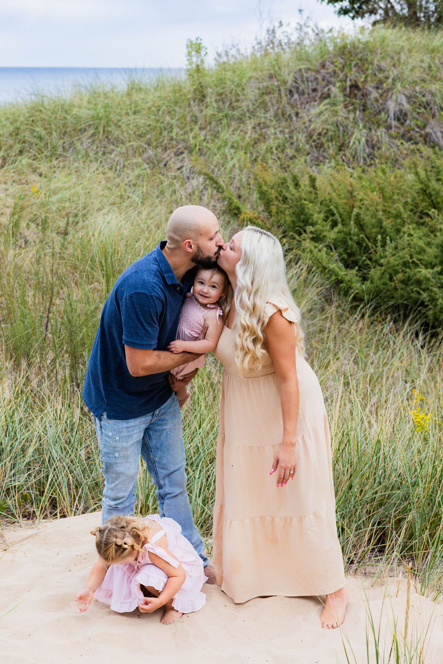 A family poses on sandy beach dunes, parents kissing their baby while a toddler plays in the sand nearby.