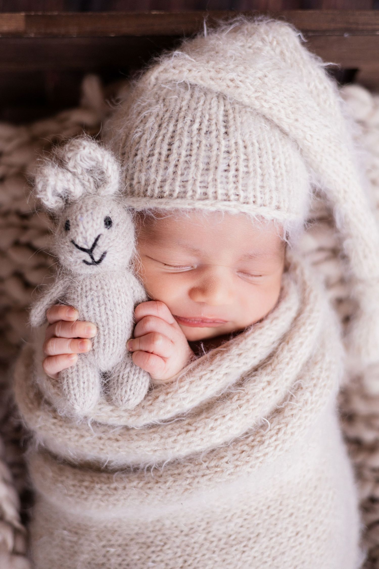 Newborn baby in cream knit wrap and tall hat, sleeping while holding a small knitted bunny toy.