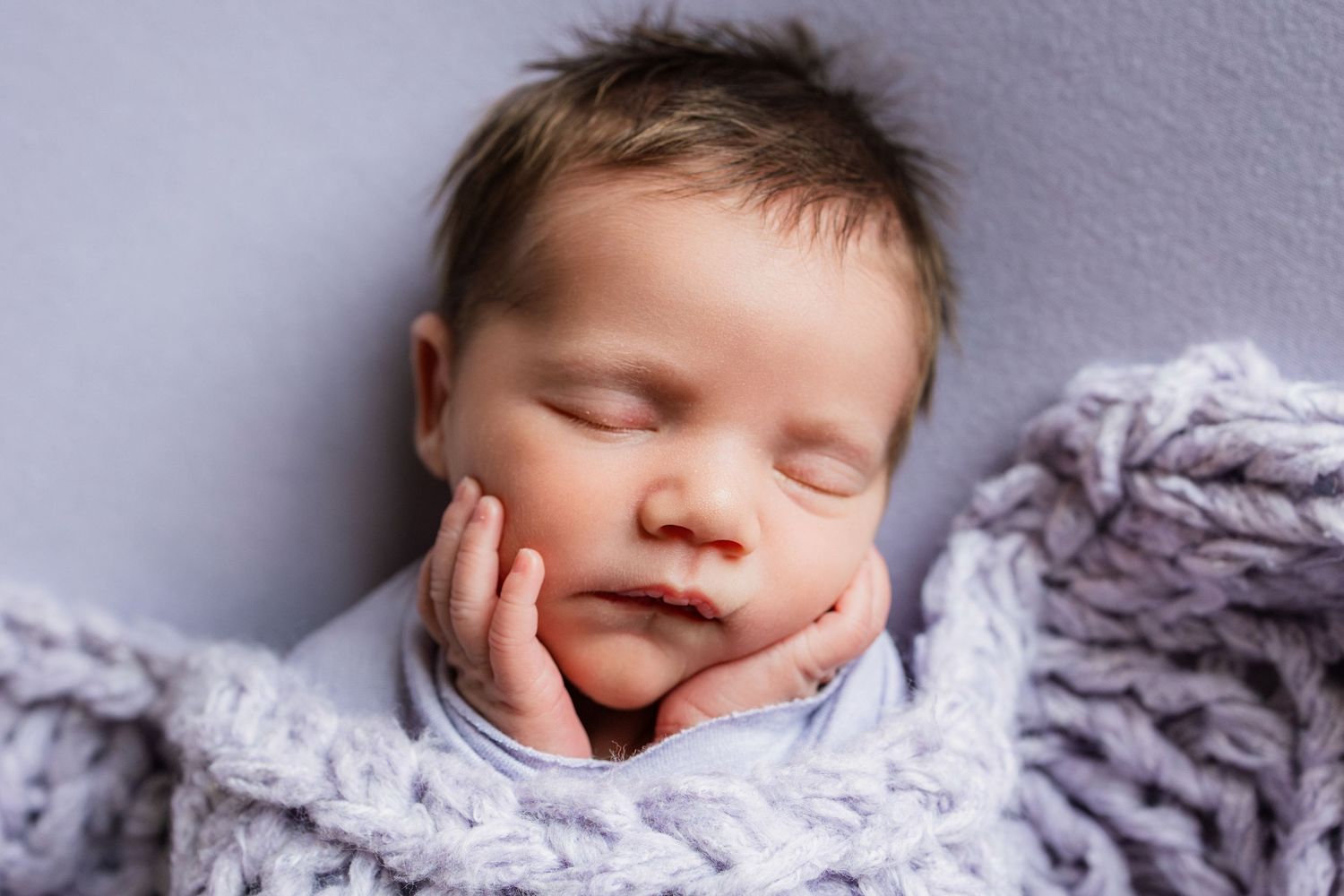 Serene newborn baby resting with tiny hands cradling face, swaddled in a cozy lavender knit wrap.