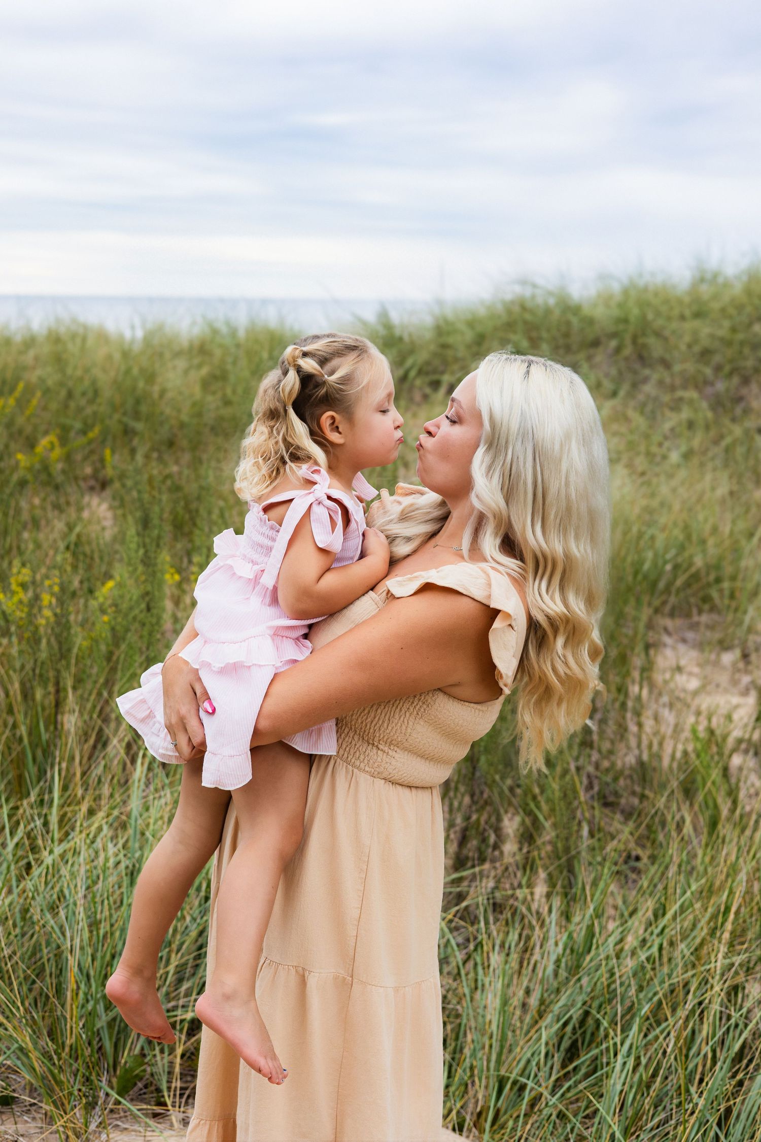 A blonde woman in a tan dress holds a young girl in a pink outfit near beach dunes.
