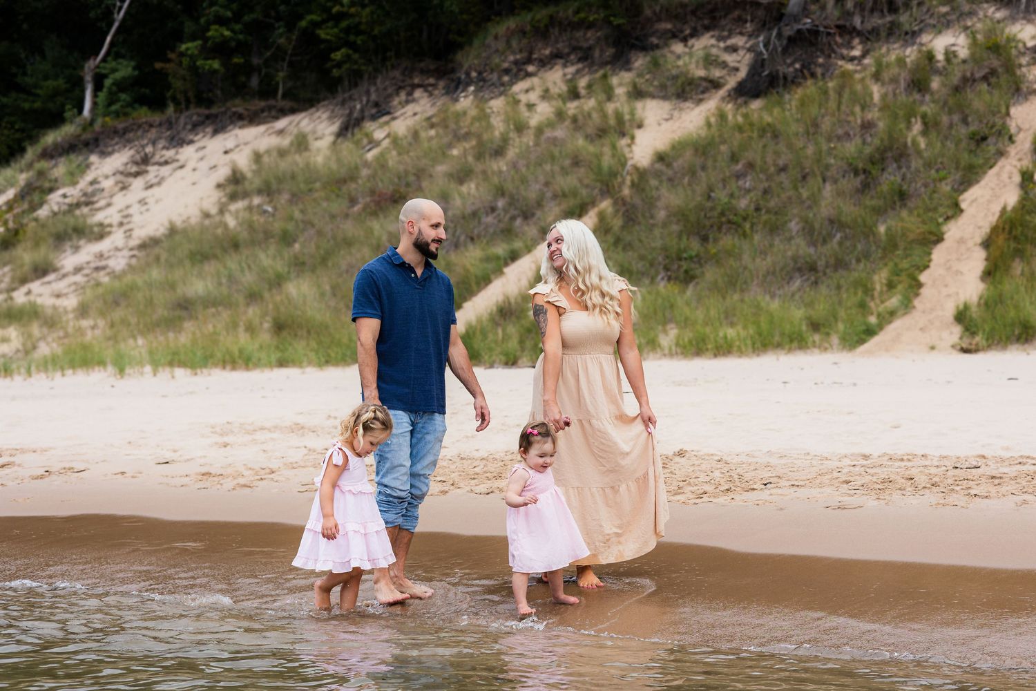 Family of four walking on sandy beach shoreline, parents with two young daughters in white dresses on sunny day.