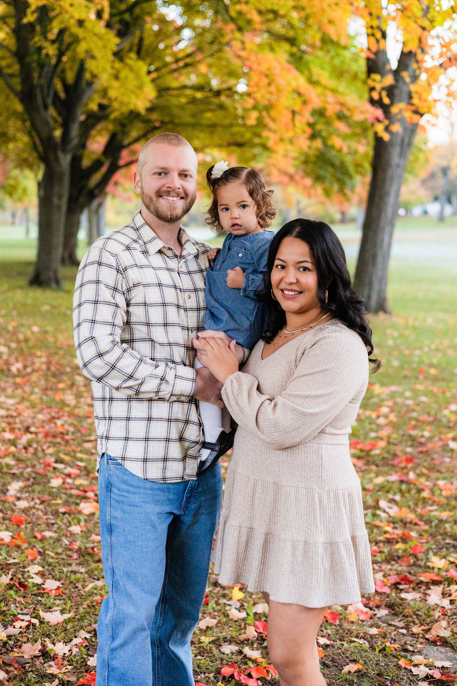 A happy family of three poses outdoors among colorful autumn trees, with dad holding their toddler and mom smiling beside them.