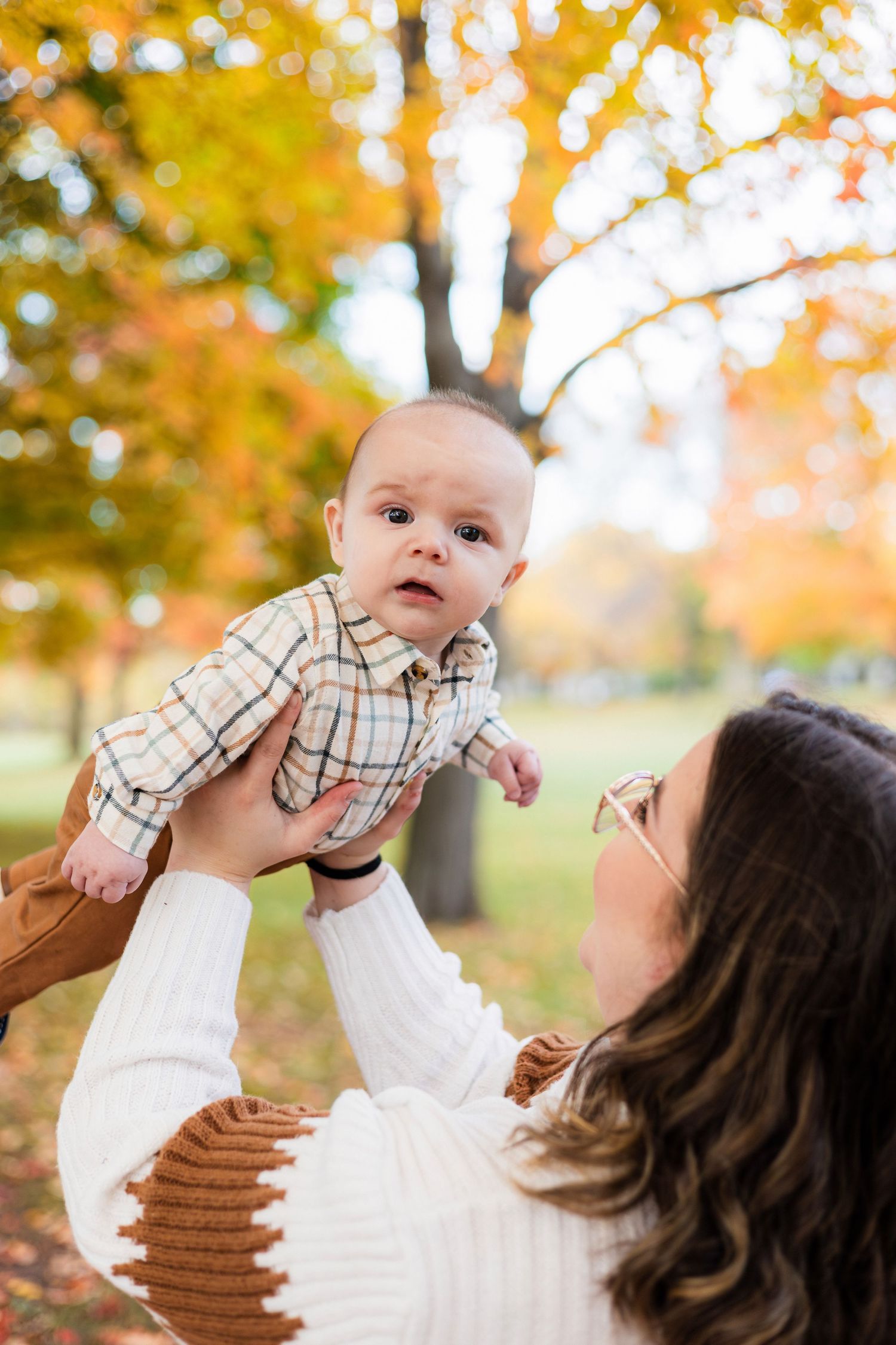 Mother in white sweater lifting curious baby in plaid shirt against colorful autumn foliage in a park setting.