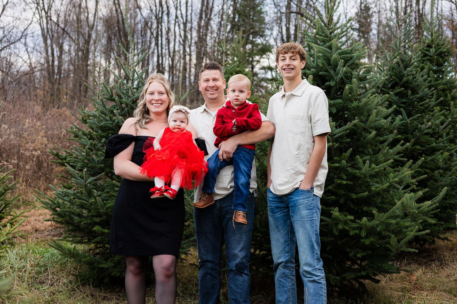 Family of five posing at a Christmas tree farm, with baby and toddler in red outfits for holiday photos.