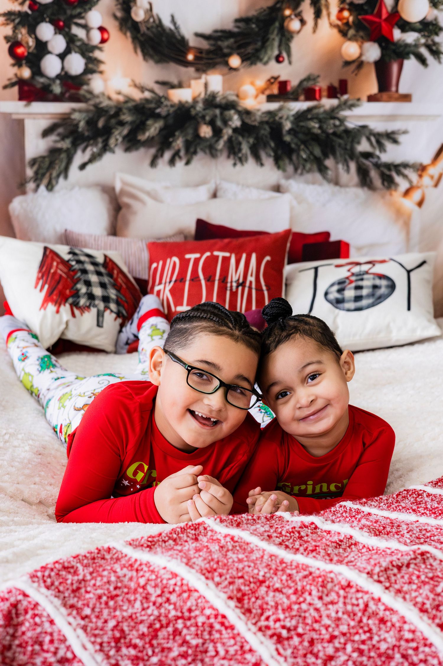 Two children in red Christmas pajamas lying on a festive decorated bed smiling together.