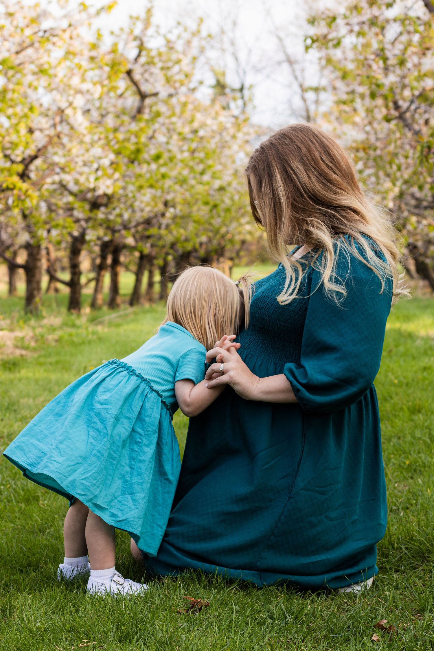 Pregnant woman in teal dress crouching as young toddler girl in blue dress hugs her baby bump in a blooming orchard.