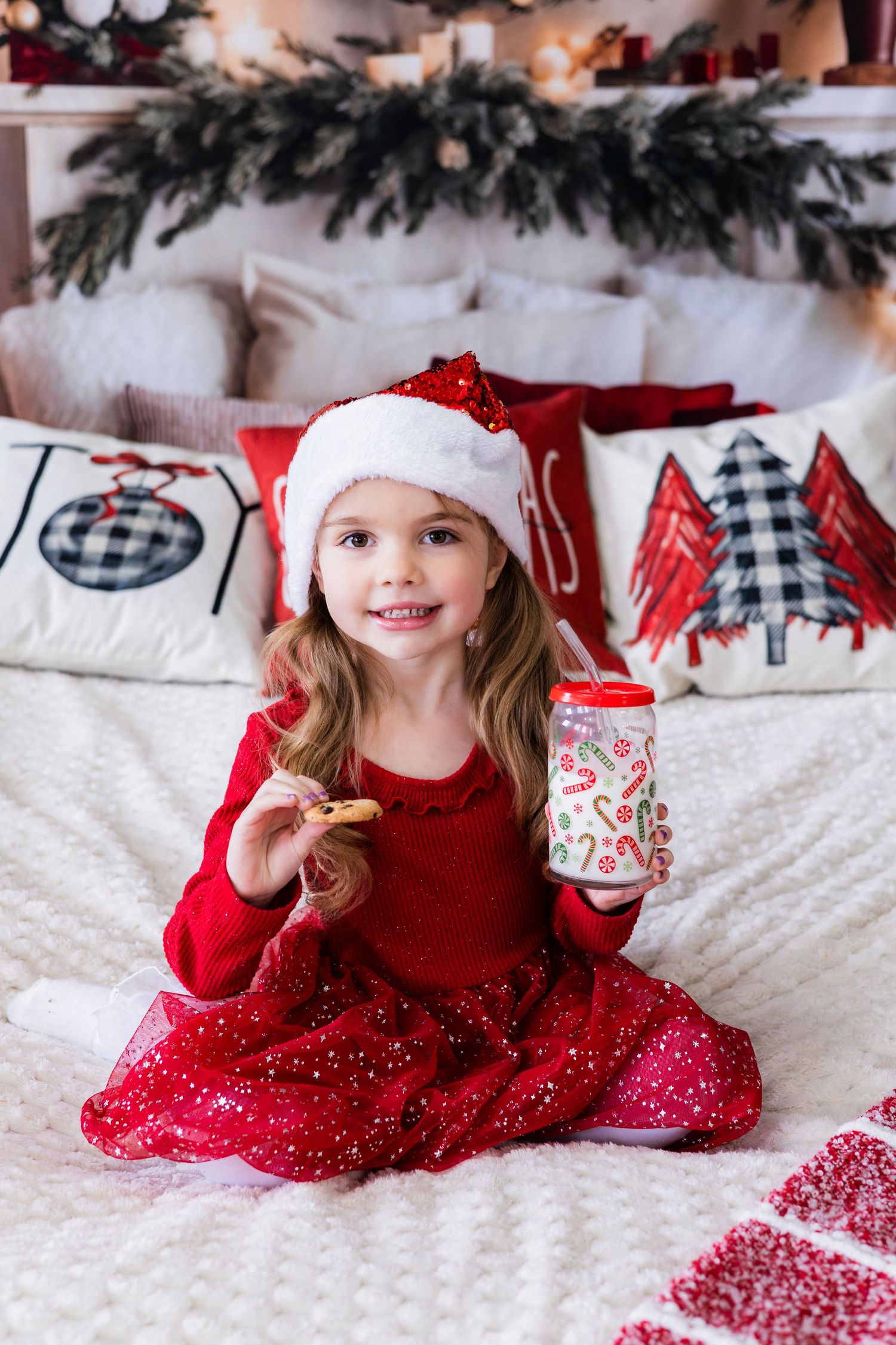 Adorable girl in red dress and Santa hat sitting on bed holding cookie and festive cup.