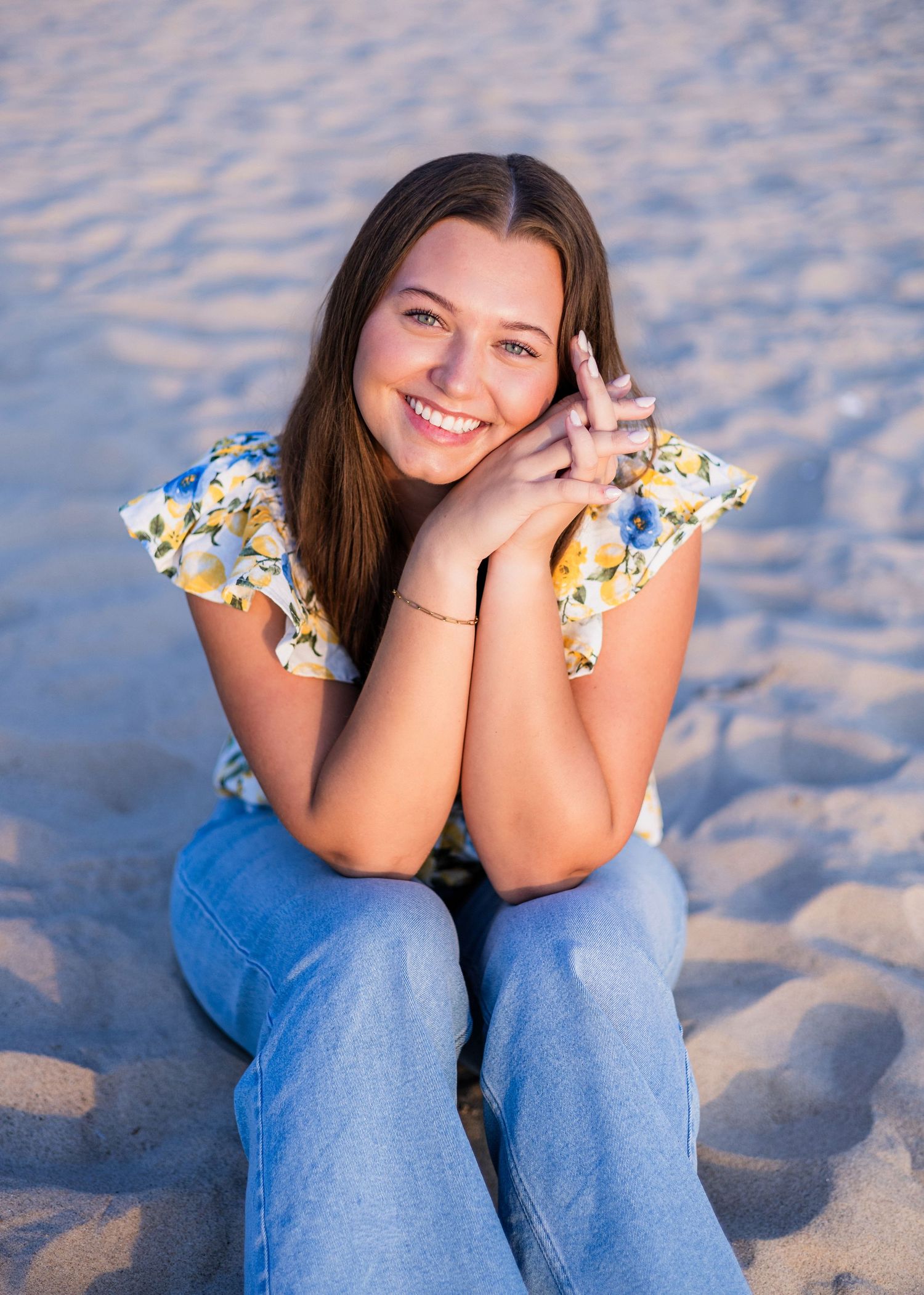 Smiling teenage girl with long brown hair sits on sandy beach wearing floral top and jeans.