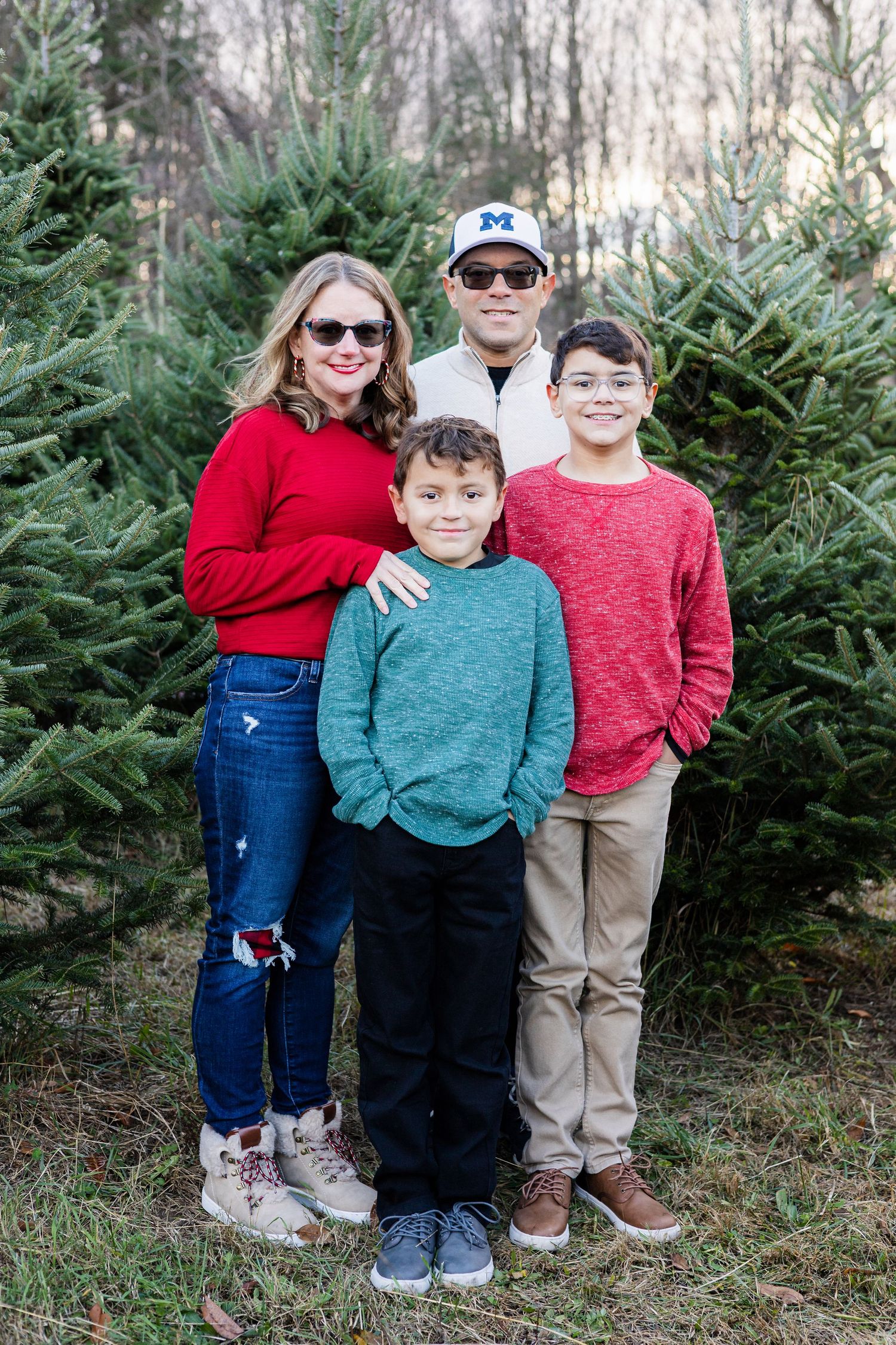 Family of four posing among Christmas trees, wearing red and teal sweaters for a festive holiday photo session.