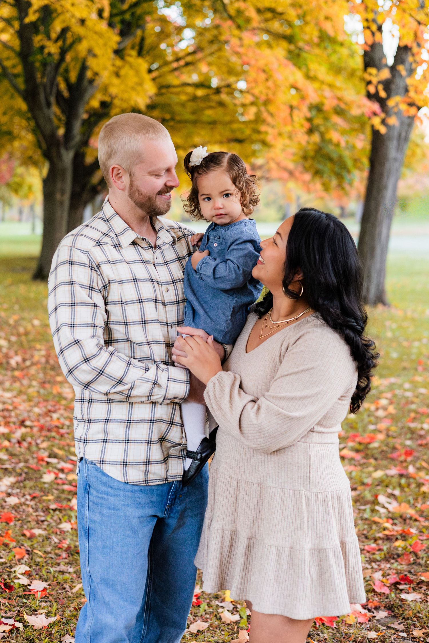 Young family with toddler girl enjoying autumn foliage in a park during a fall family photo session.